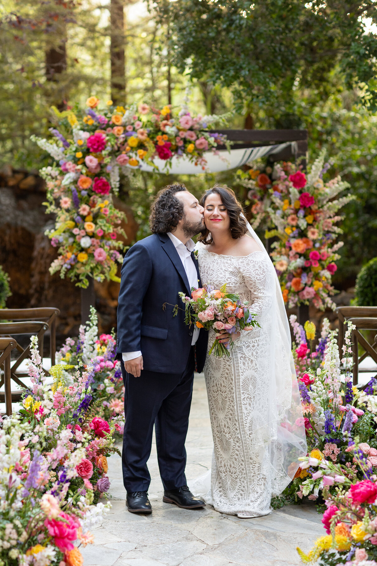 A groom kissing a bride's cheek while they are surrounded by flowers