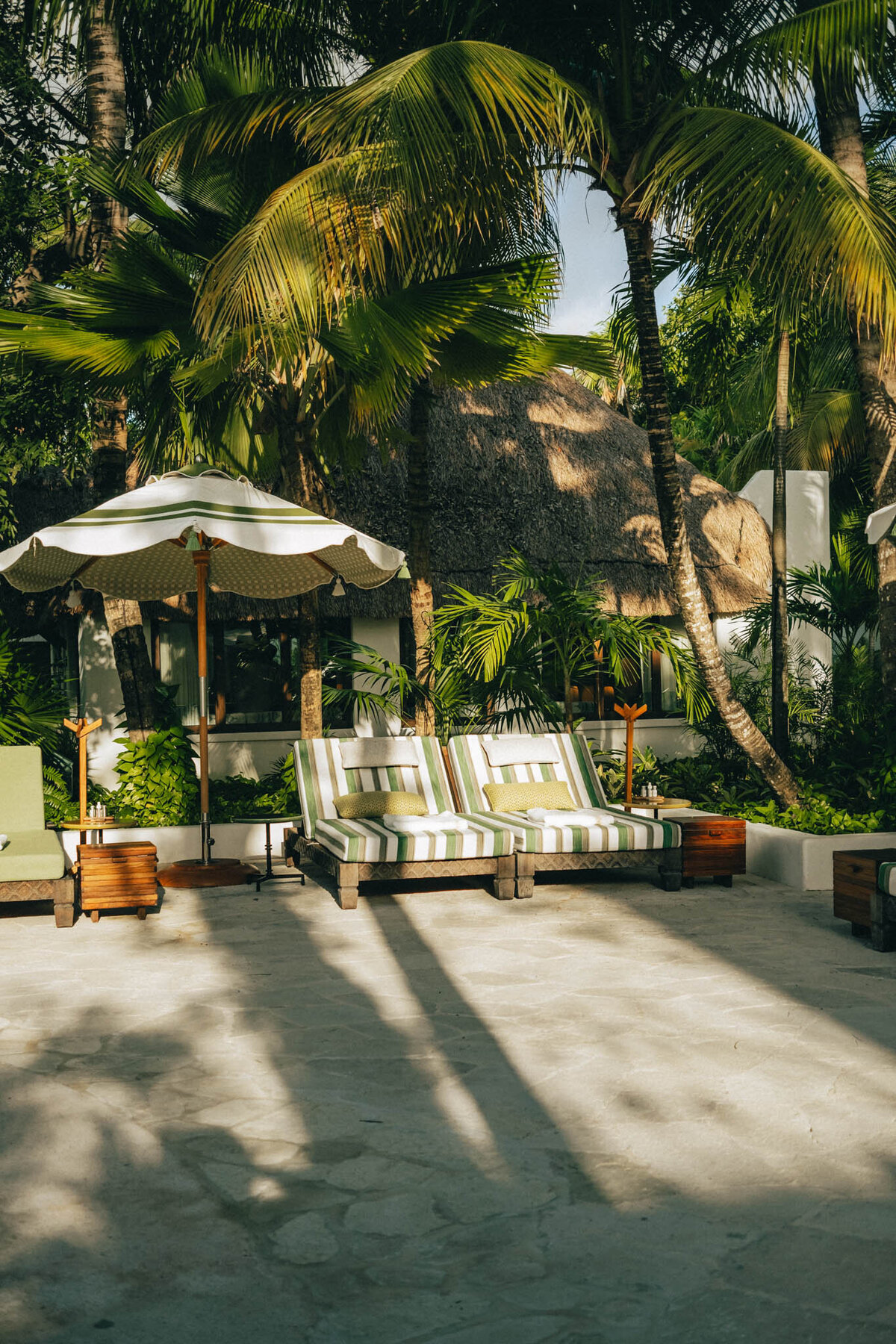 Lounge area with two striped sunbeds and a large white umbrella surrounded by lush tropical greenery and tall palm trees. A thatched-roof structure is visible in the background under a clear blue sky.