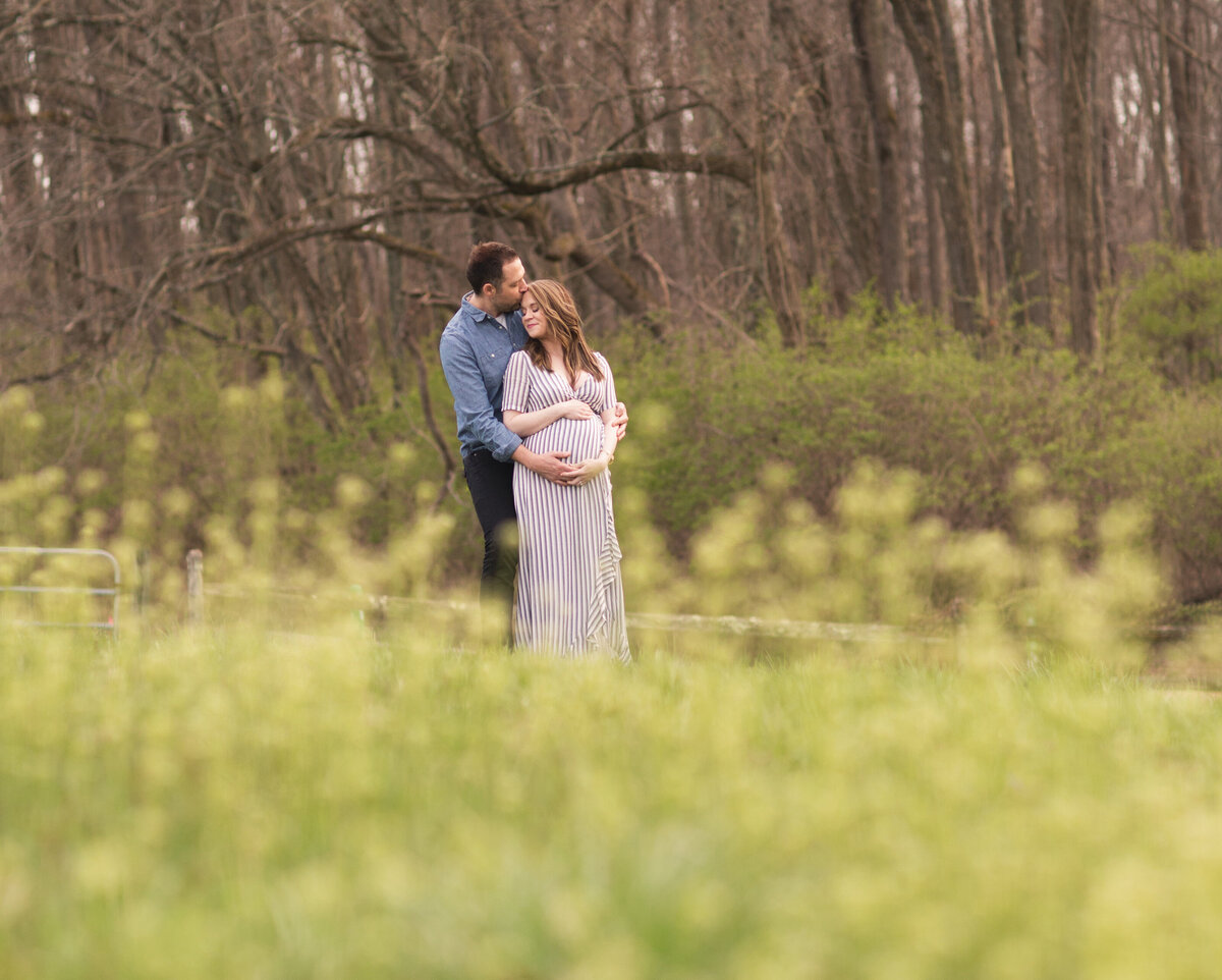 Outdoor maternity photo of a pregnant wife and her husband standing together in a sunlit field