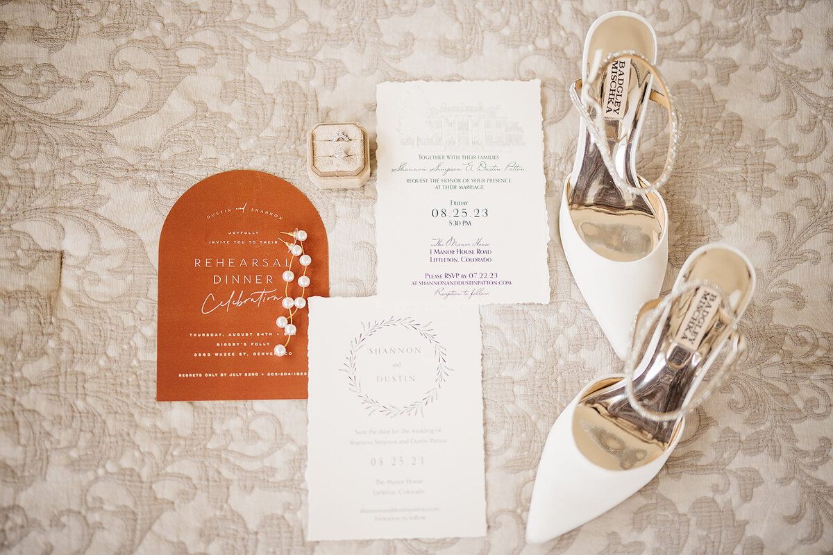 Wedding day details with an orange invitation, white detail pages, and a white pair of heels.