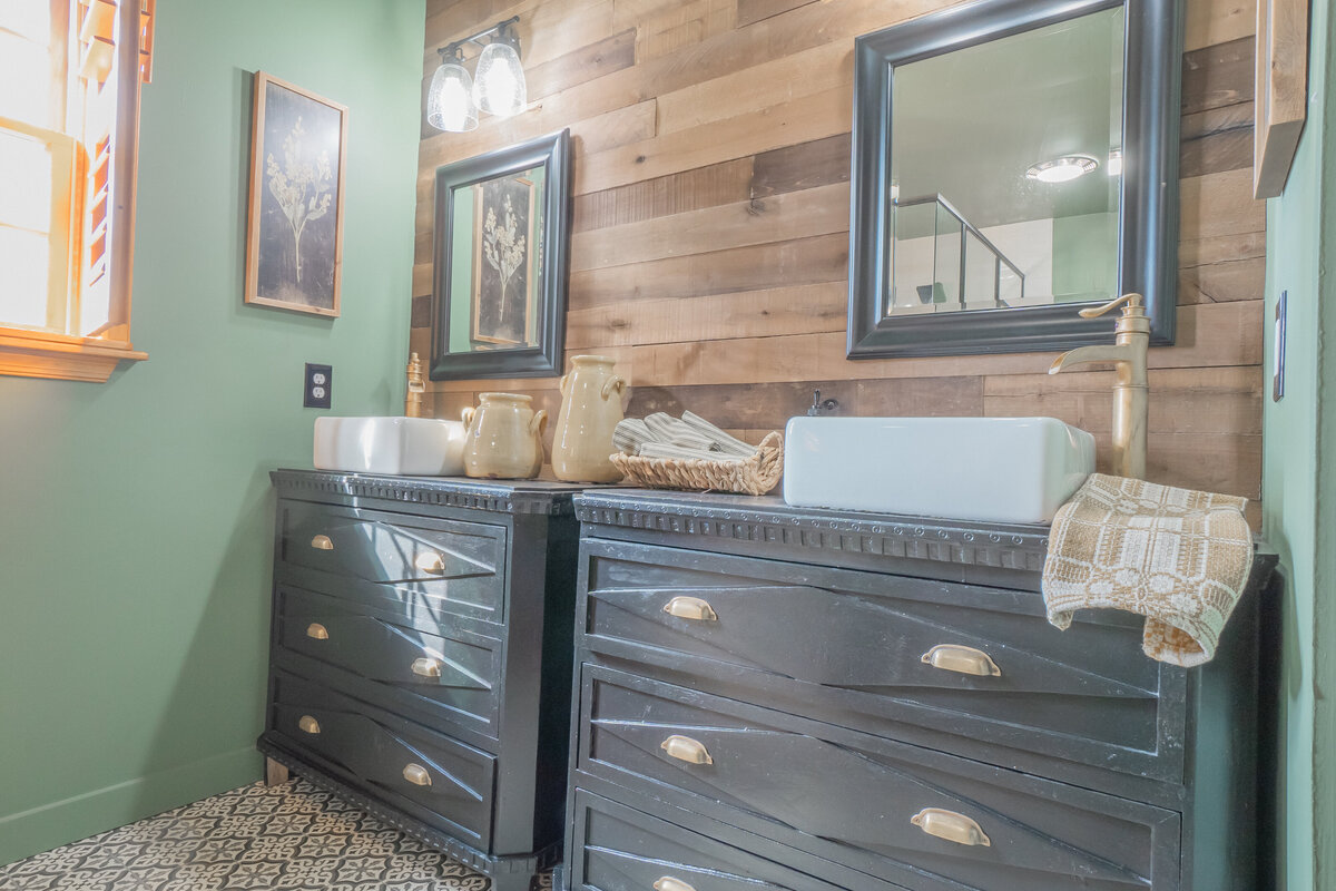 Wooden slat wall with black framed mirrors and black cabinets with brass cup handles with white sink vessels