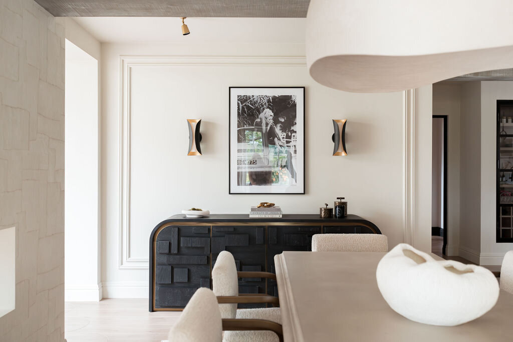 A modern dining room features a large light-colored table with beige padded chairs. On the wall, a black sideboard with a photograph above it is flanked by two contemporary sconces. The space is minimalistic with neutral tones and a sculptural white centerpiece on the table.