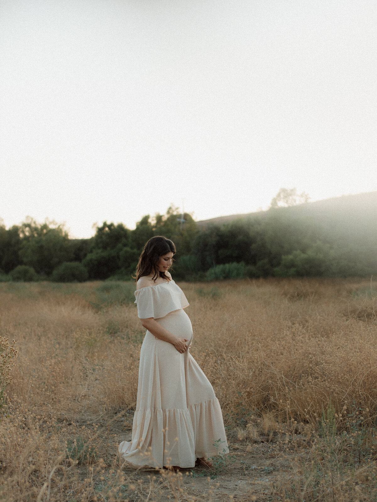 Pregnant mother standing in a golden field at sunset, tenderly holding her belly in a flowing cream dress.