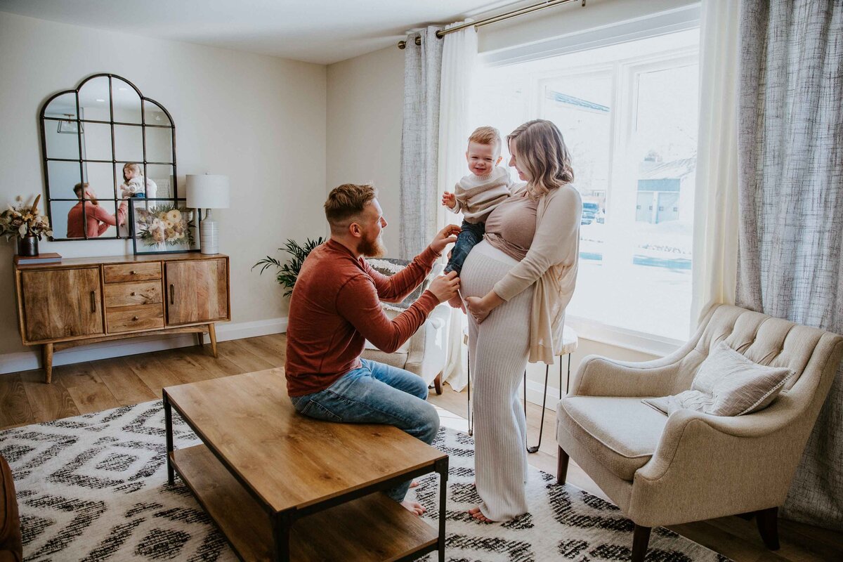 In home maternity session in London, Ontario. Expectant mom and dad are standing with their toddler in living room. Mom is holding toddler, dad is sitting on the coffee table tickling the toddler.