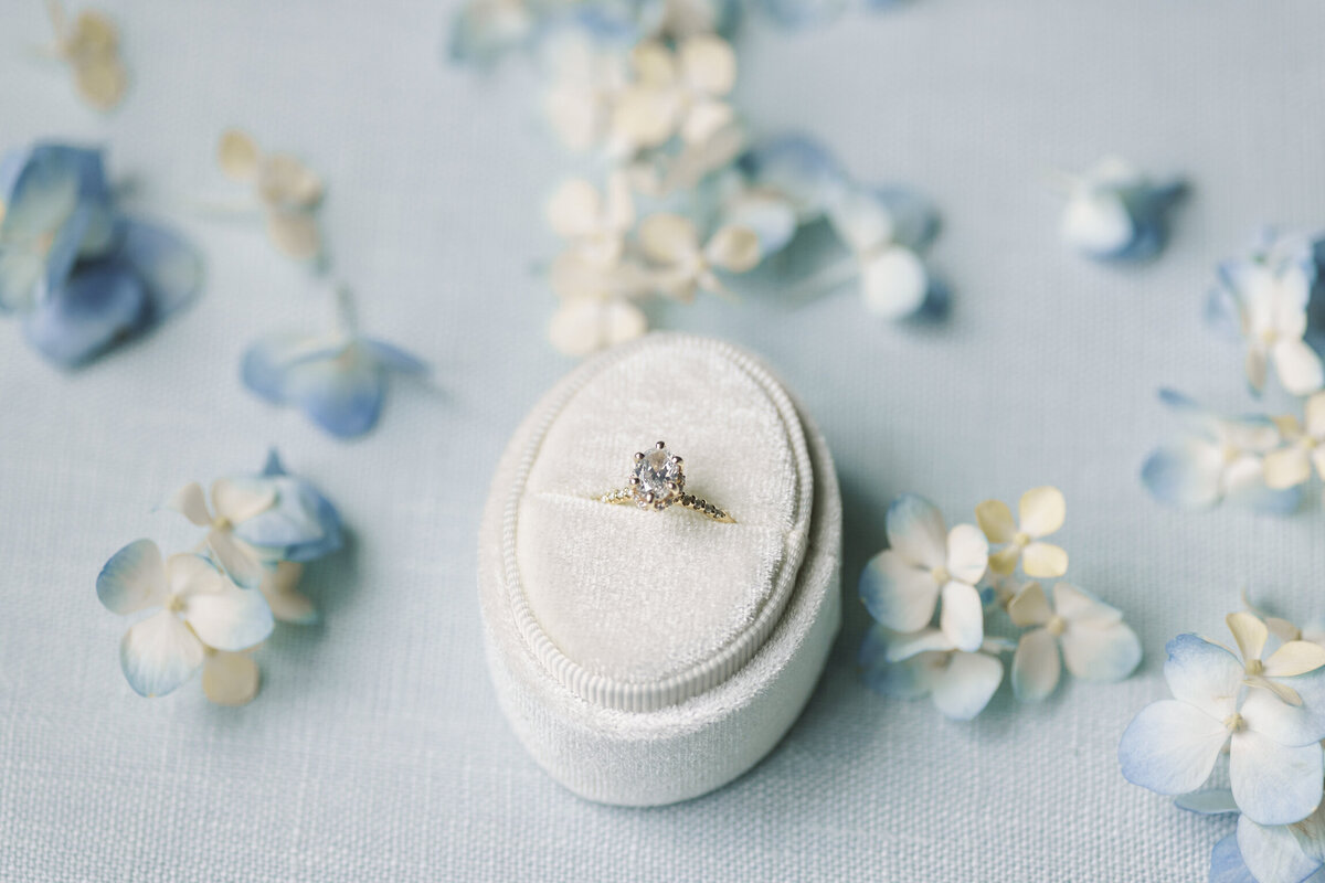 A stunning engagement ring displayed against a soft blue background, accented by delicate blue and white hydrangeas, artfully captured on film by Morgan Alysse, Louisiana film photographer.