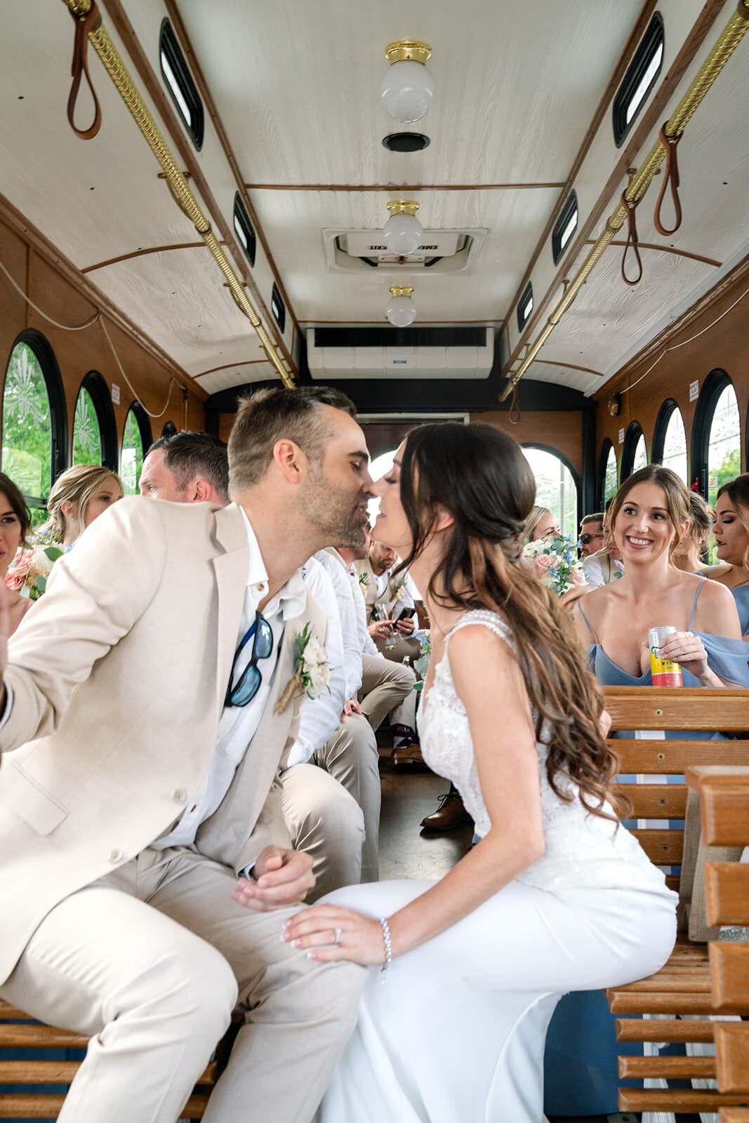 Inside the trolley, the bride and groom sit facing each other, holding hands, and sharing a kiss, surrounded by their bridal party.