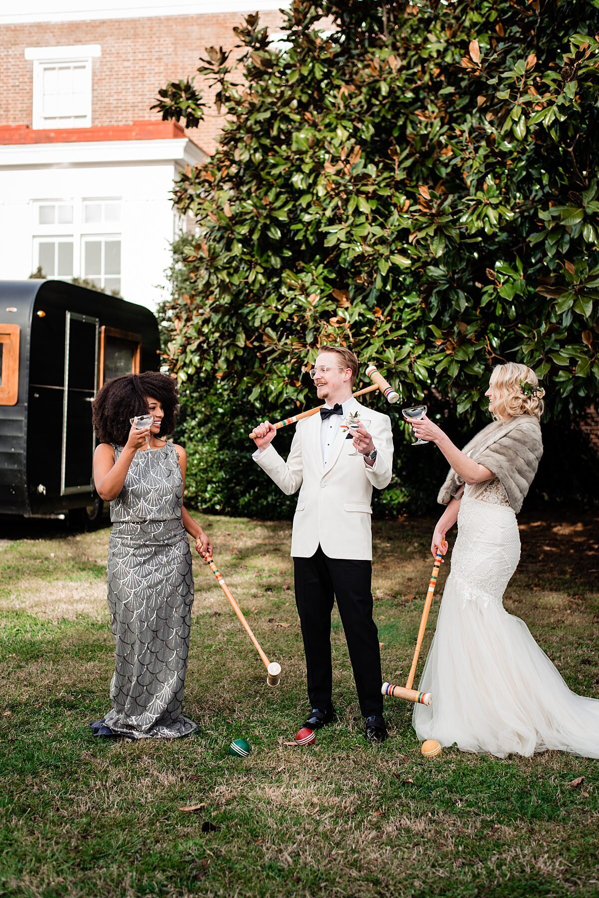 African American bridesmaid wearing a silver sheath dress plays croquet with the groom wearing a white jacket and black pants and the 1920s bride wearing a long white lace dress and fur wrap while drinking champagne