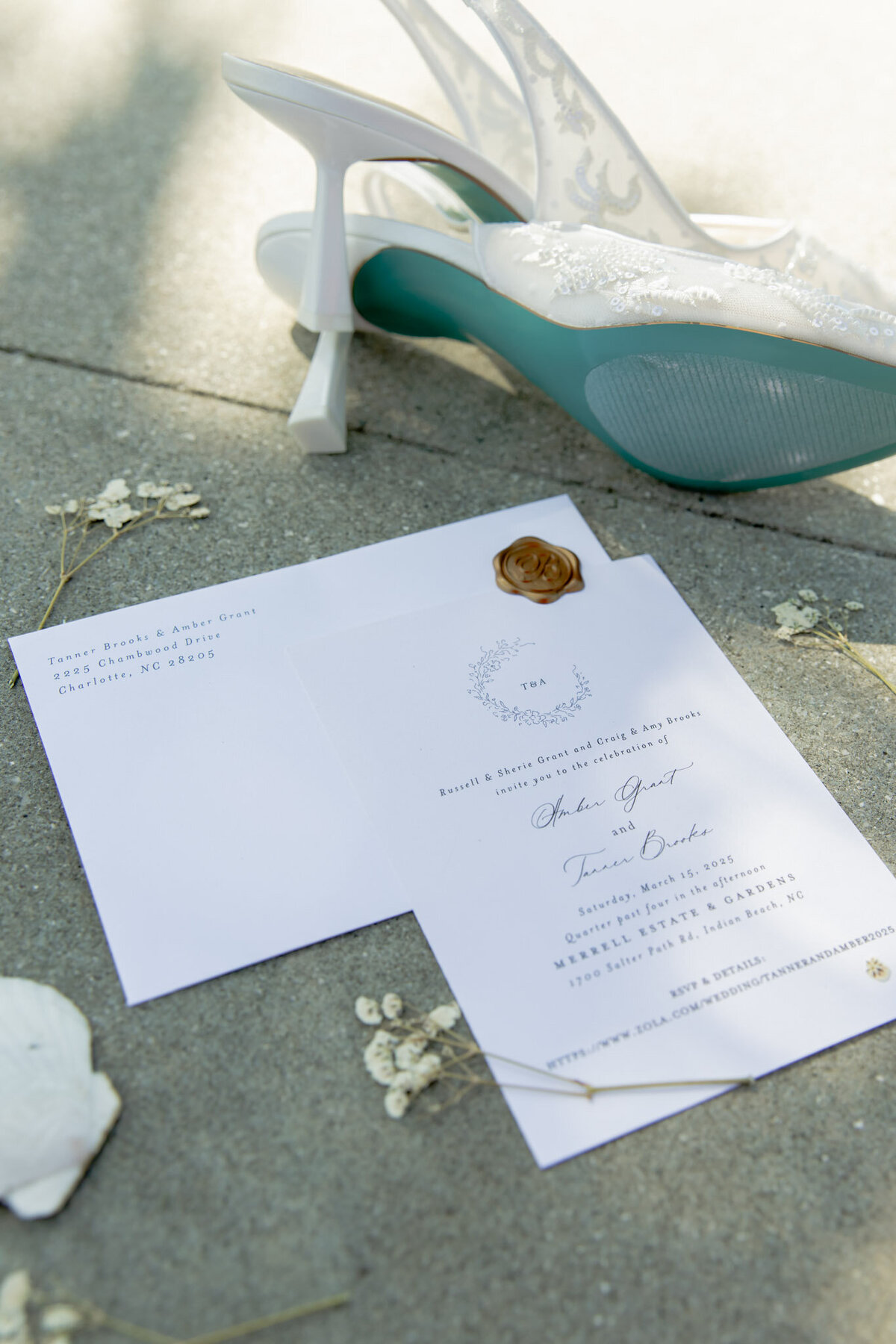 A pair of white high heels, a wedding invitation, and a few small white flowers are laid on the concrete ground.