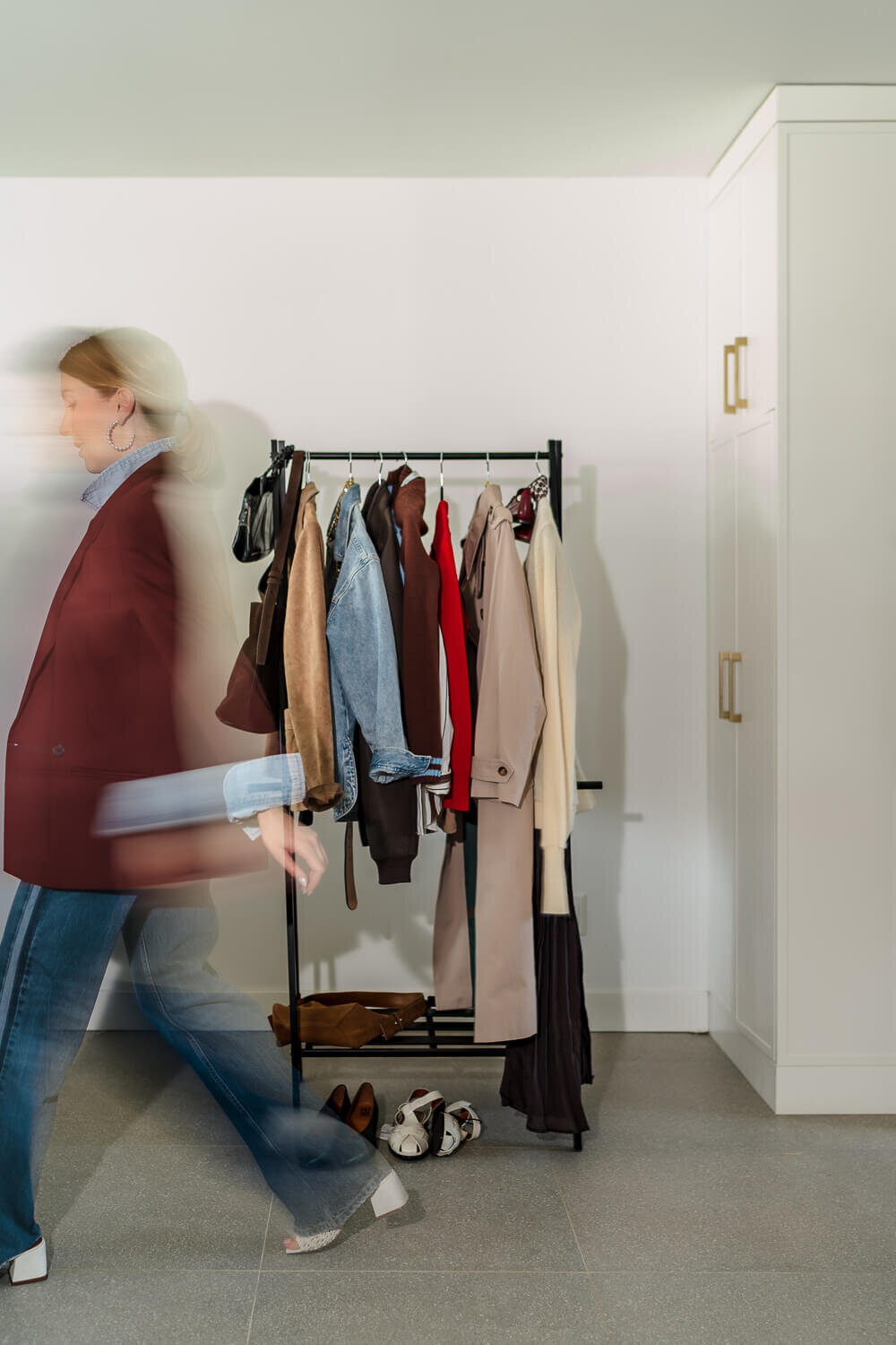 Motion-blurred woman in maroon blazer walking past clothing rack with assorted jackets and coats.
