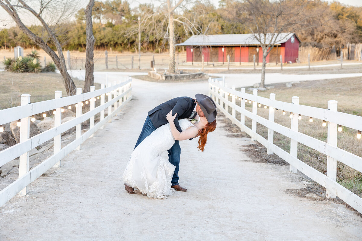 groom dips bride in kiss between white fence lines and red barn behind in Boerne Texas