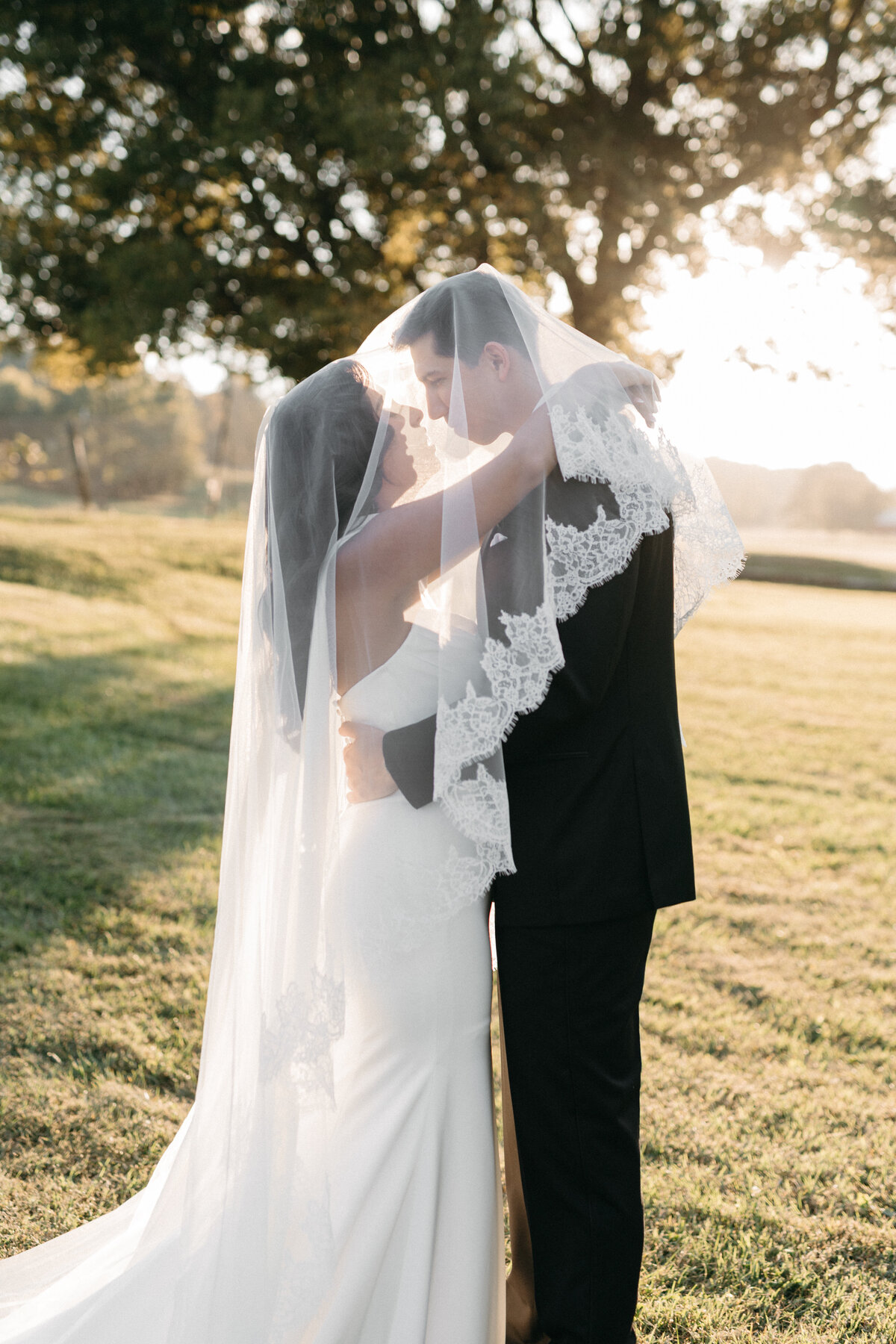 Golden-hour portrait of the bride and groom embracing under a delicate lace-edged veil, highlighting soft, romantic wedding florals and sunset light for dreamy outdoor wedding inspiration.
