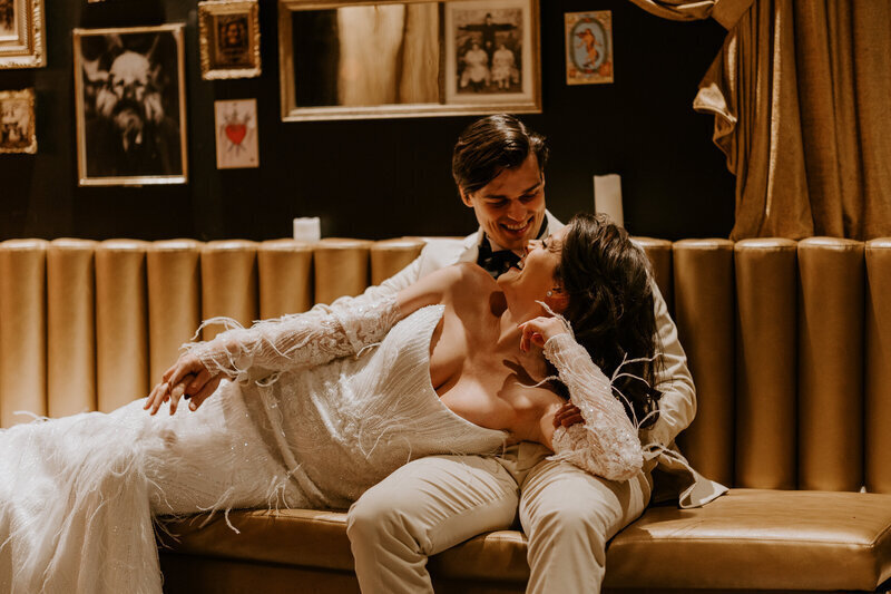 Mariée allongée sur les genoux du marié, assis sur un canapé vintage camel et se regardant dans les yeux. Pausent pour Laura, photographe de mariage professionnelle.