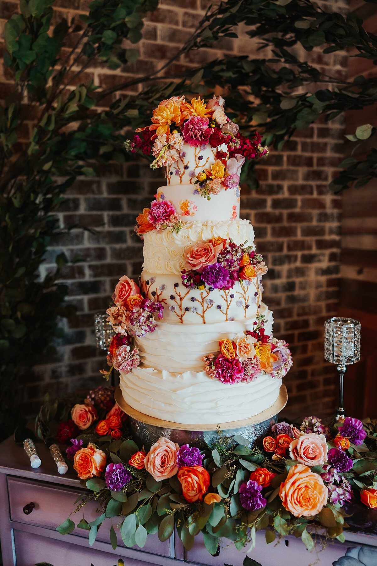 Very tall seven tier wedding cake with clusters of peach roses, pink roses, purple roses and orange roses sitting on a purple dresser with a large assortment of roses and a crystal candle holder around the base. There is a brick wall and arching ficus tree behind the cake at Steel Magnolia Barn at a Nashville Wedding.