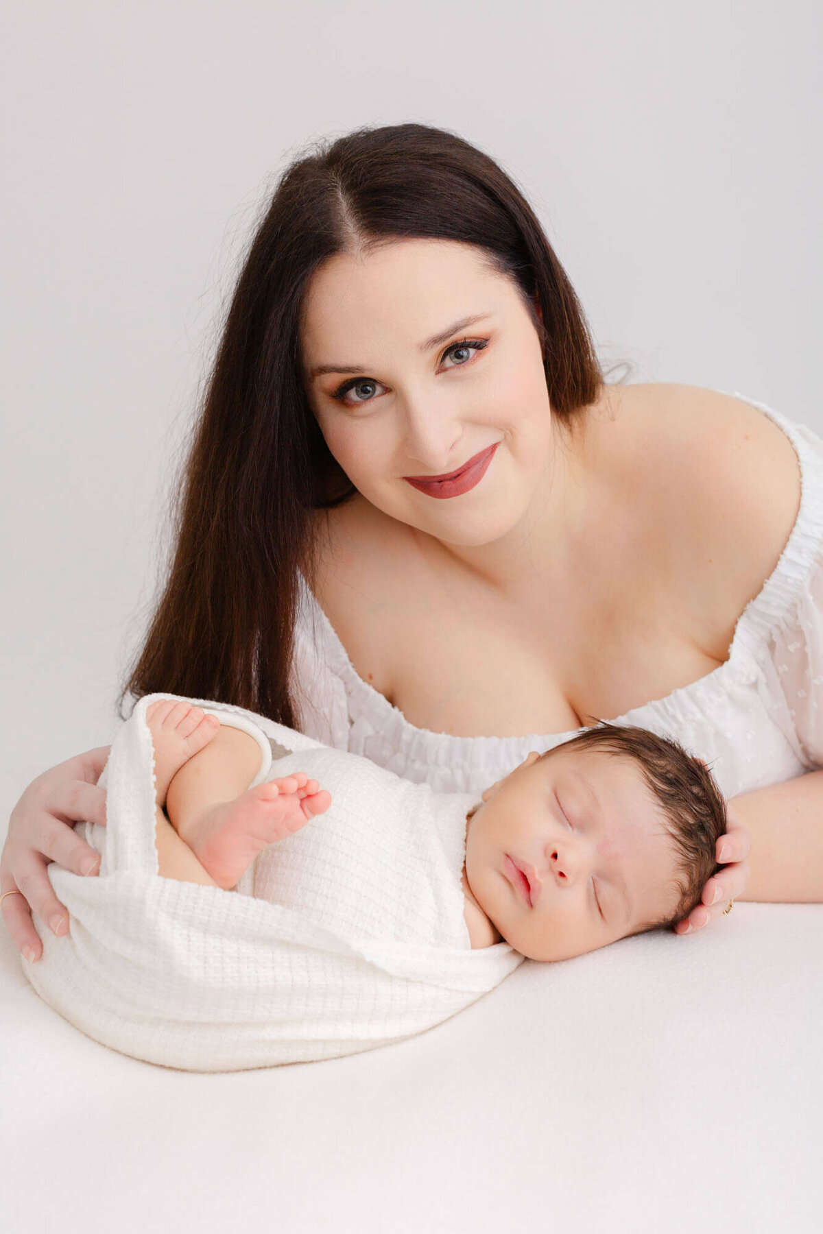 Mom holding her baby while baby is resting on a white blanket. Mom is in an off the shoulder white dress and she is looking up and smiling at the camera during baby's newborn photoshoot in PDX.