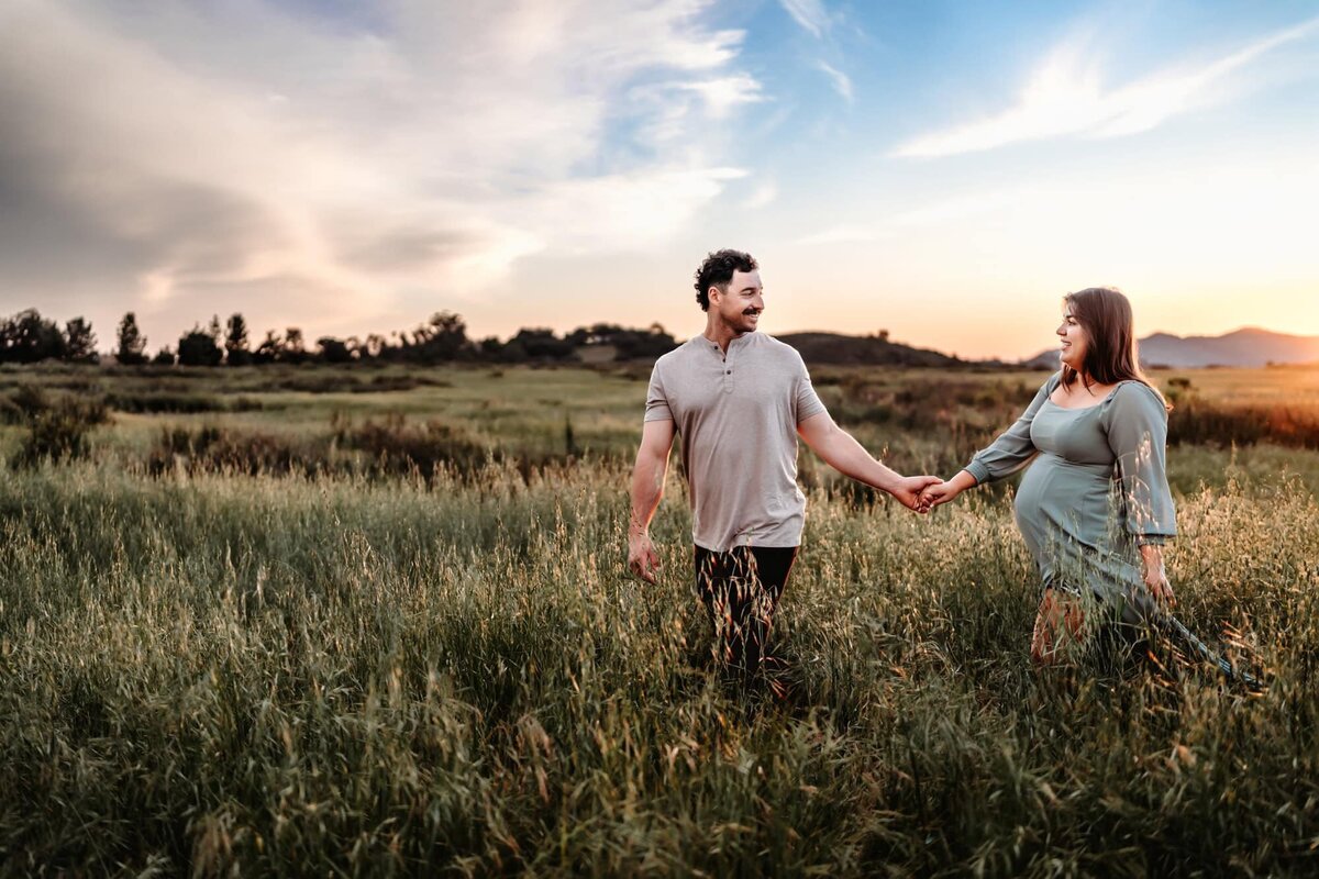 A man hold his pregnant wife's hand, leading her thorugh a field of tall grass during their maternity photoshoot in San Diego. 