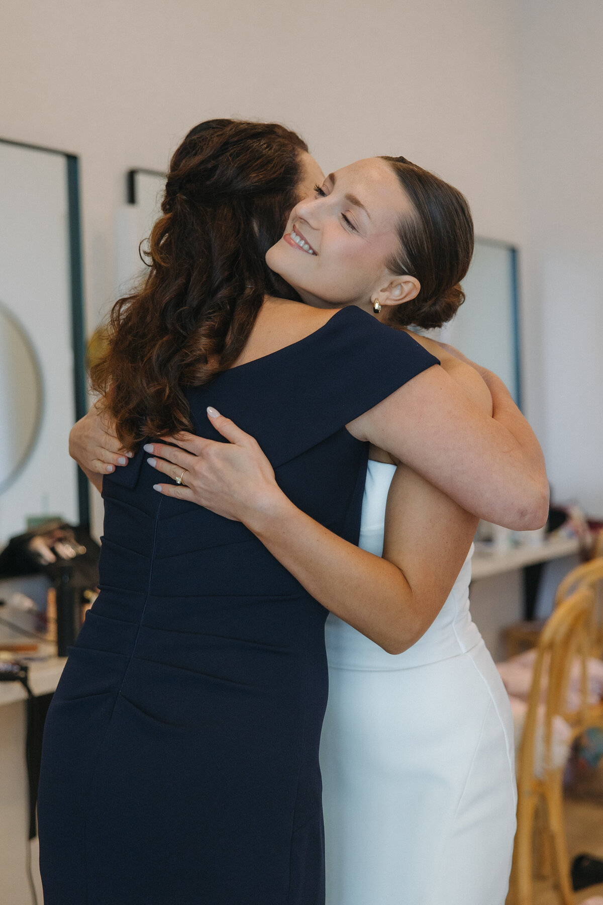 A bride hugs another women in a formal navy blue dress. The bride is smiling into the woman's shoulder. Her hair is in a slicked back bun and she has small gold hoop earrings in. Her dress is fitted to her body and white. 
