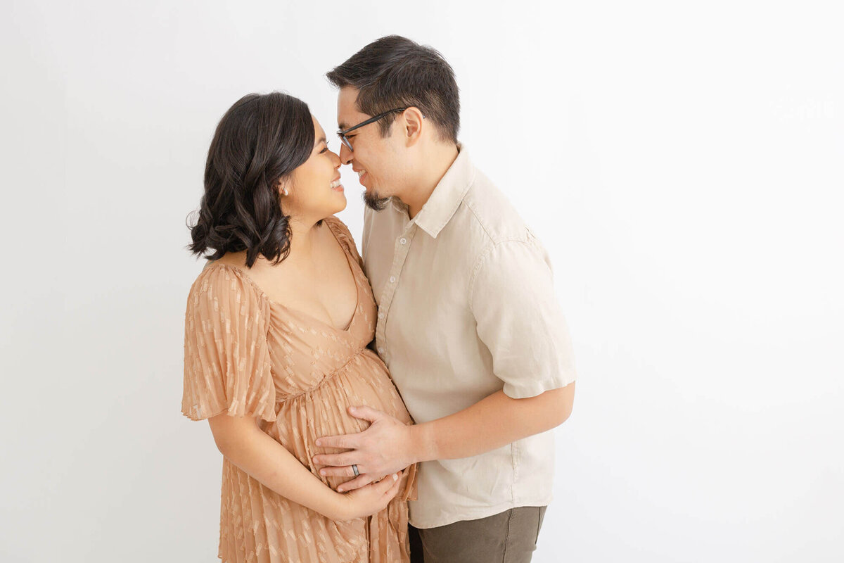 Parents to be, standing nose to nose and laughing with one another while they both hold onto Mama's baby bump.  They are in a Portland Maternity Photographer's all-white portrait studio in SE Portland. Photo by Ashlie Behm Photography