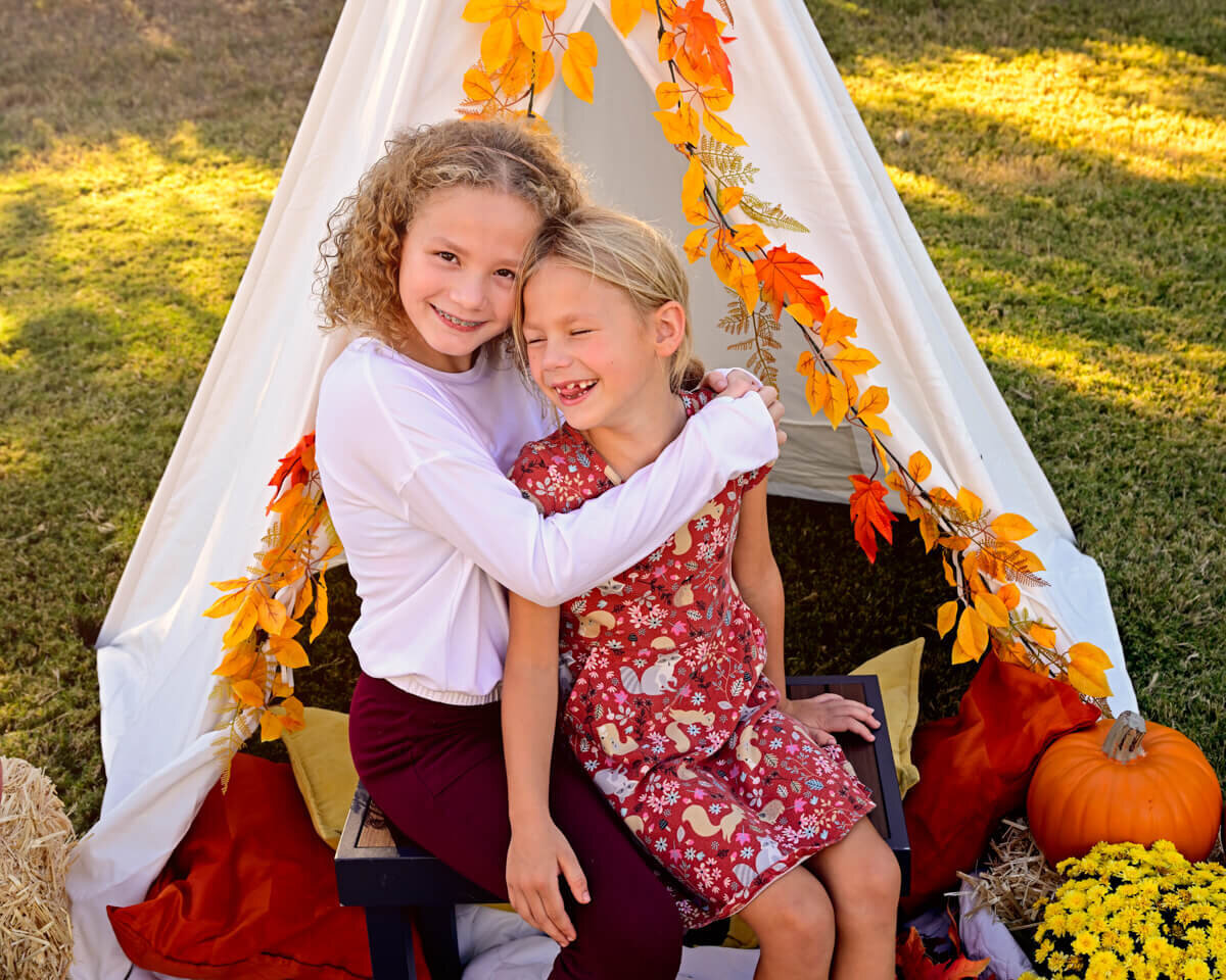 McKinney, TX outdoor family photography session of two young sisters laughing in their tent decorated with fall colored leaves.