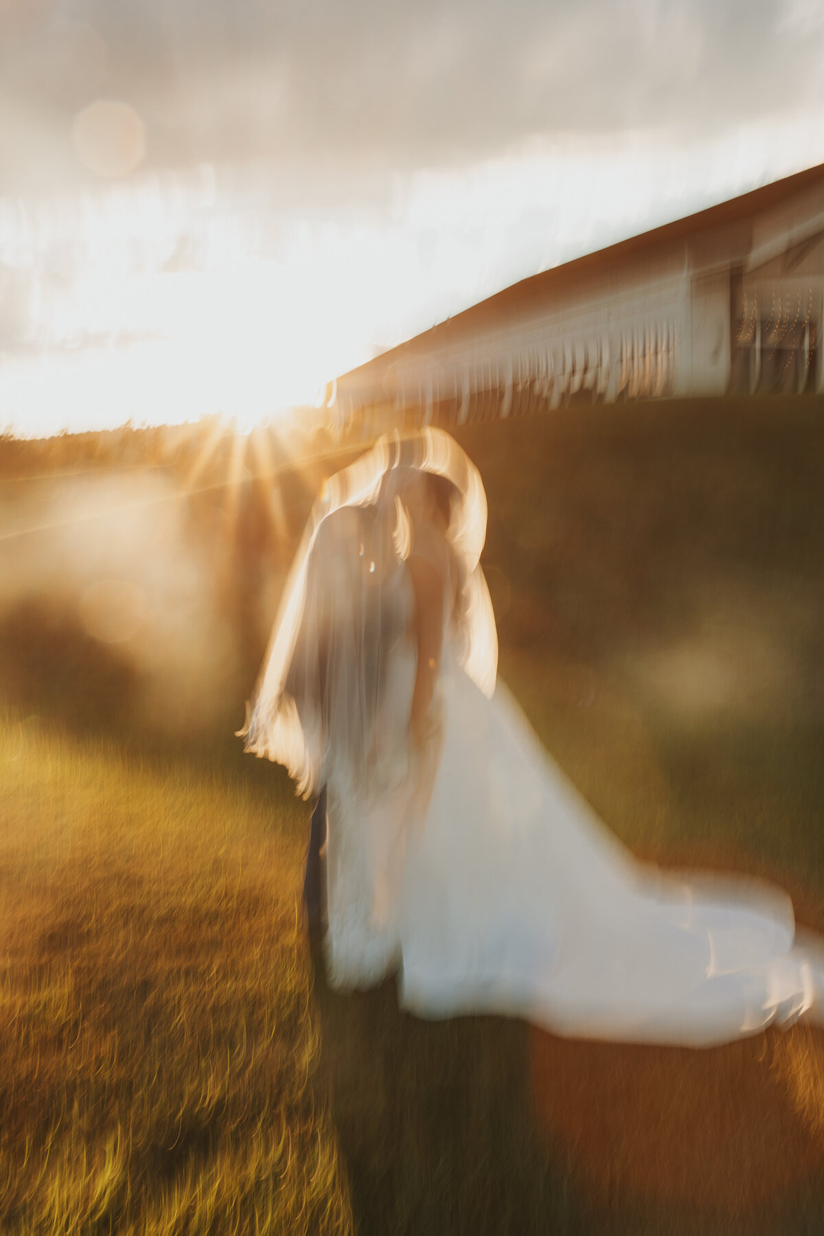 Blurry photo of bride and groom kissing as the sun sets behind them.