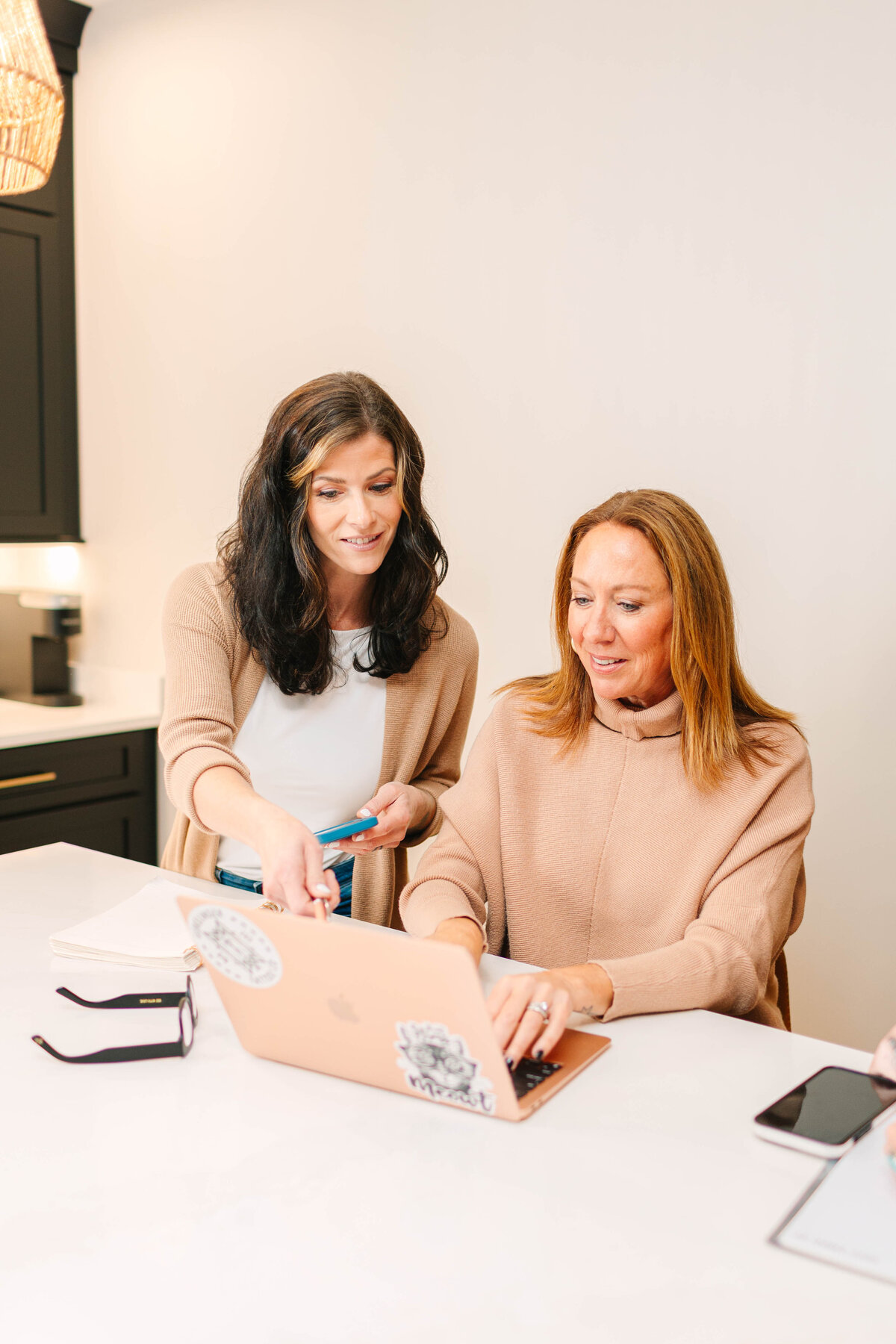 Two professional women collaborate at a white kitchen island. One, holding a smartphone, points at the screen of a rose gold laptop, while the other types. They are dressed in neutral tones, with notebooks and glasses on the table, and a modern kitchen in the background.