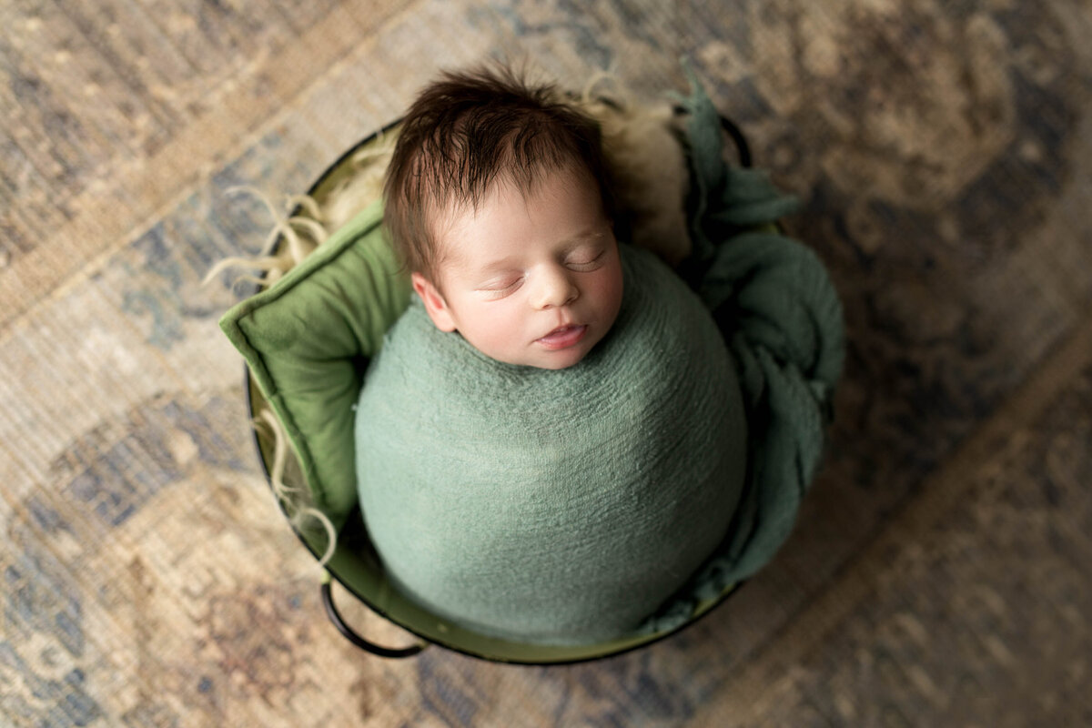 Studio newborn photography - baby sleeping in basket wrapped in a green knit swaddle on top of a vintage Persian patterned rug.
