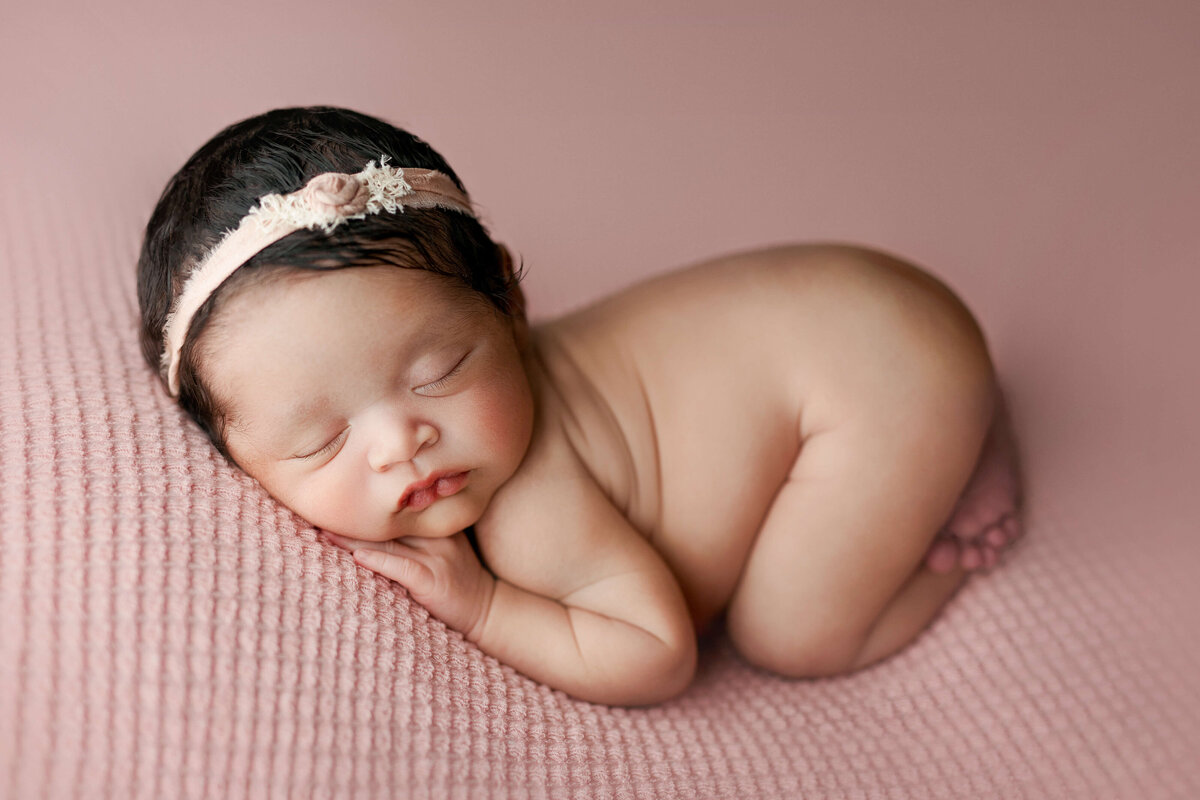 Newborn girl laying on her tummy  on a pink backdrop with matching headband at an in-studio  newborn photo shoot in south riding va