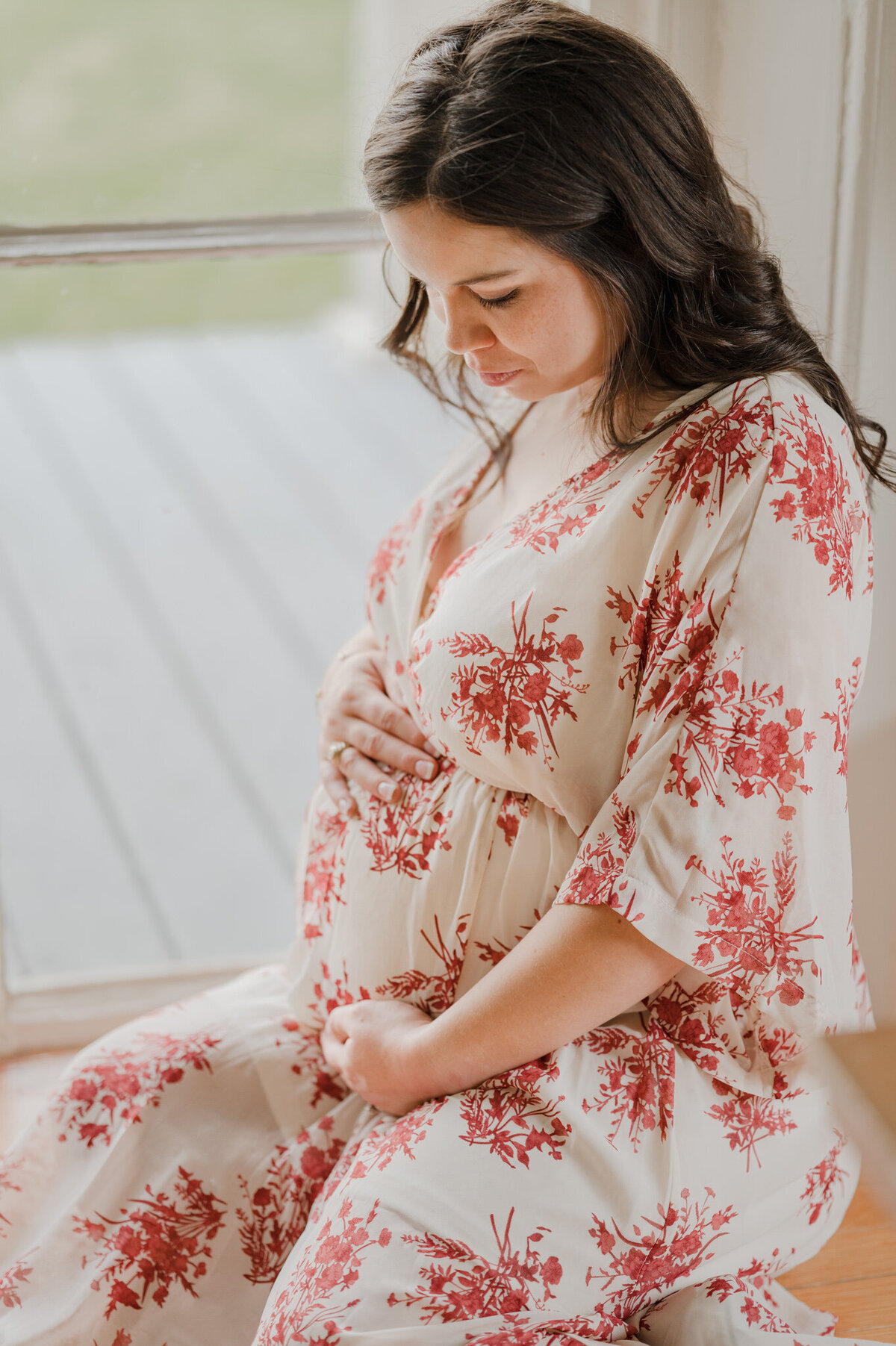 Expecting woman in a gown sits inside a window and cradles her bump.