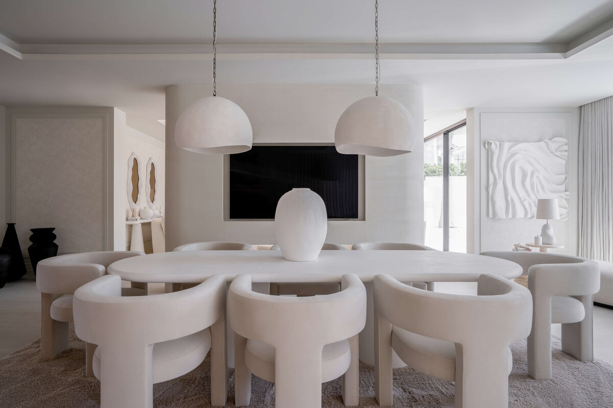 A modern dining room with a minimalist aesthetic, featuring a large oval table surrounded by six curved white chairs. Two large, white dome-shaped pendant lights hang above the table. The room is decorated in neutral tones with a plush, light-colored rug underneath the table and chairs. Behind the table, a large television recessed in the cream wall.