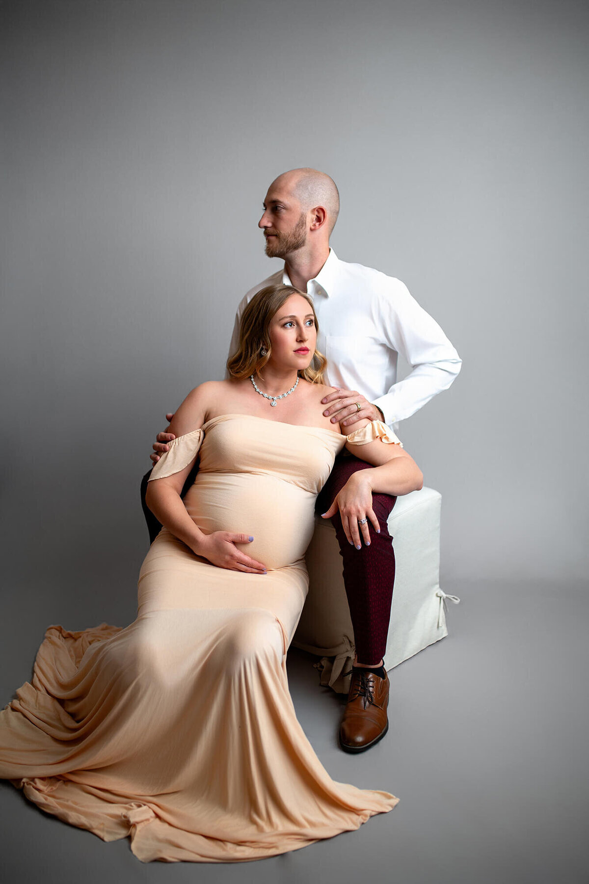 woman sitting between mans legs leaning against him while looking away in modern maternity studio session 