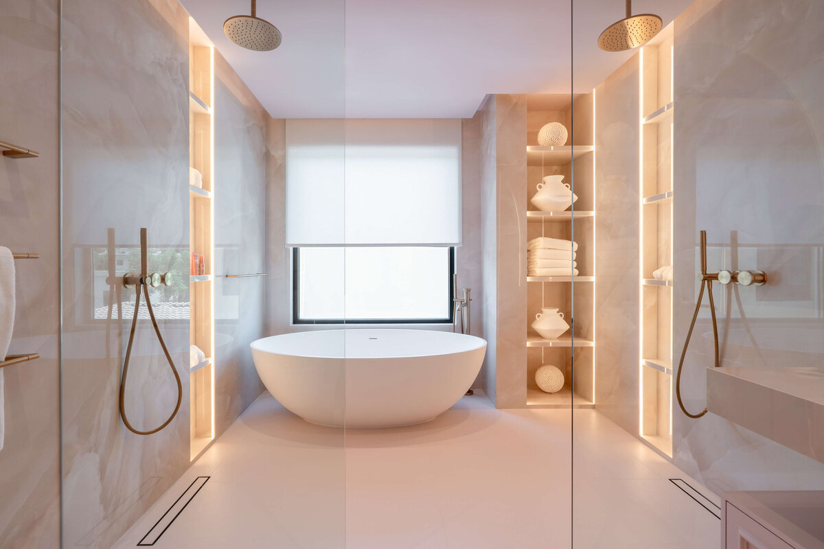 Spacious en suite bathroom with a  chic light pink aesthetic. The walls are a light pink marble. There is a large soaking tub against the far wall, and open showers with rose gold hardware on both the left and right.