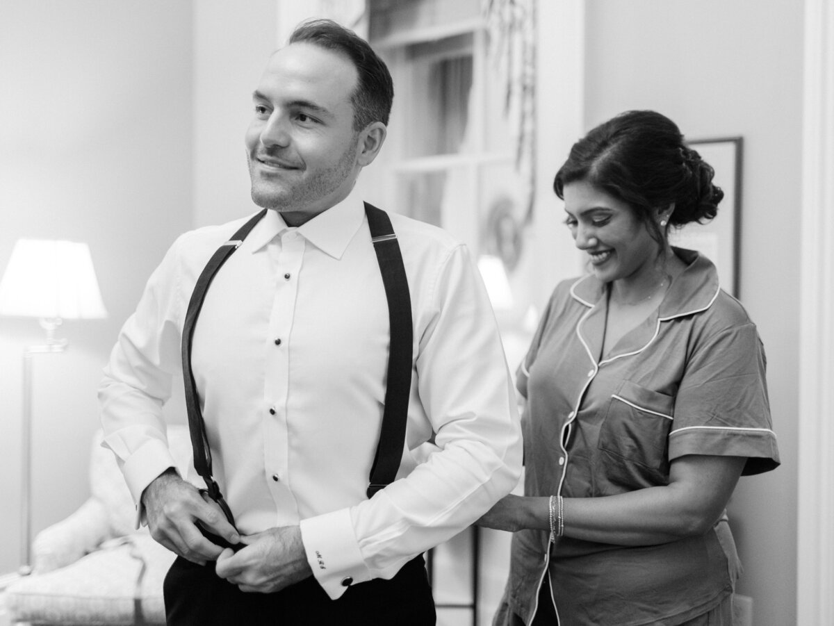 A groom getting ready, adjusting his suspenders and bowtie while a bride is assisting him, showcasing love and support in a intimate wedding moment filled with anticipation.