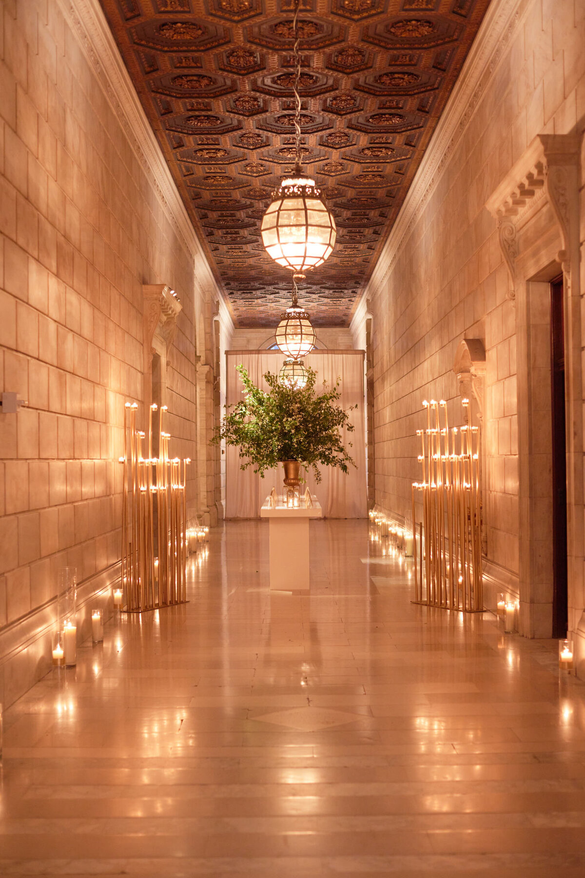 The photo was taken in a dimly lit, elegant hallway with a high, ornately designed ceiling at The New York Public Library in New York City by photographer Shawn Connell. The walls and floor are made of polished stone, creating a grand atmosphere. Tall, thin candles line the walls, casting a warm glow and adding to the ambiance. The hallway leads to a large floral arrangement at the end, beautifully illuminated by hanging lanterns, creating a striking focal point in the scene. Videographers Aaron Navak Films captured the stunning setting on video to further document the ambiance of the event.