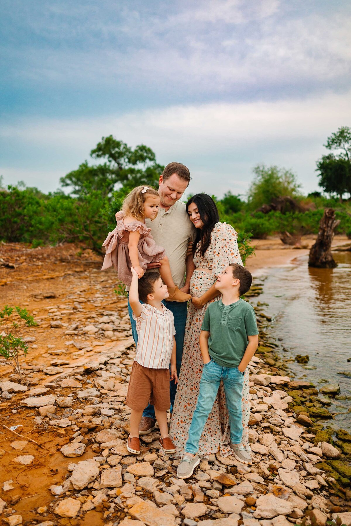 In front of a picturesque lake, the professional maternity photographer captures a joyful family moment. The parents, celebrating the arrival of their new family member, are playfully engaging with their little kids, creating a heartwarming and vibrant scene.