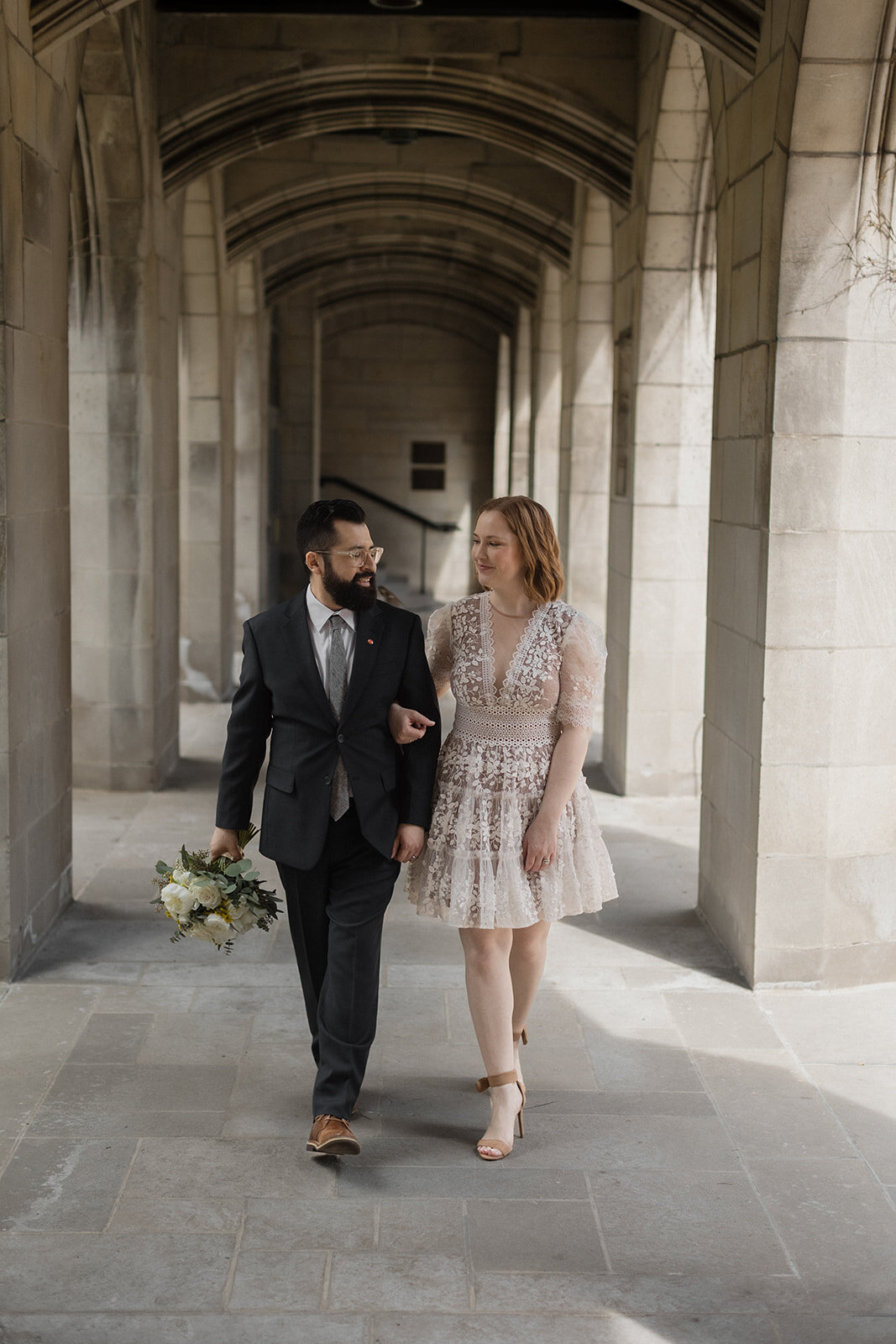 Wedding couple walking in a cloister with beautiful light streaming through. Her arms is linked in his and he carries the bouquet and they look at one another.