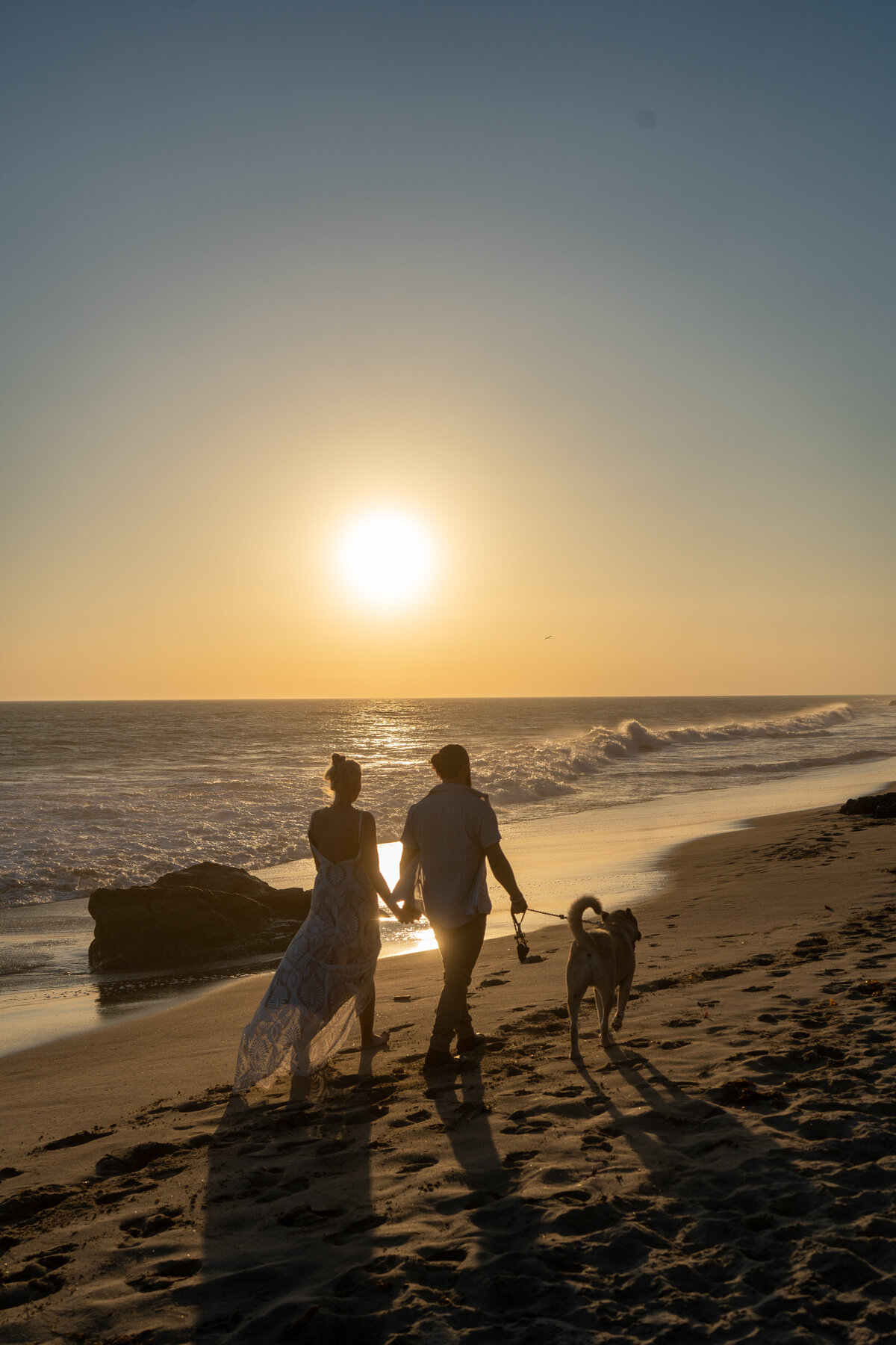 Silhouettes of the bride, groom, and dog at the beach in Leo Carillo, Malibu walking near the waves