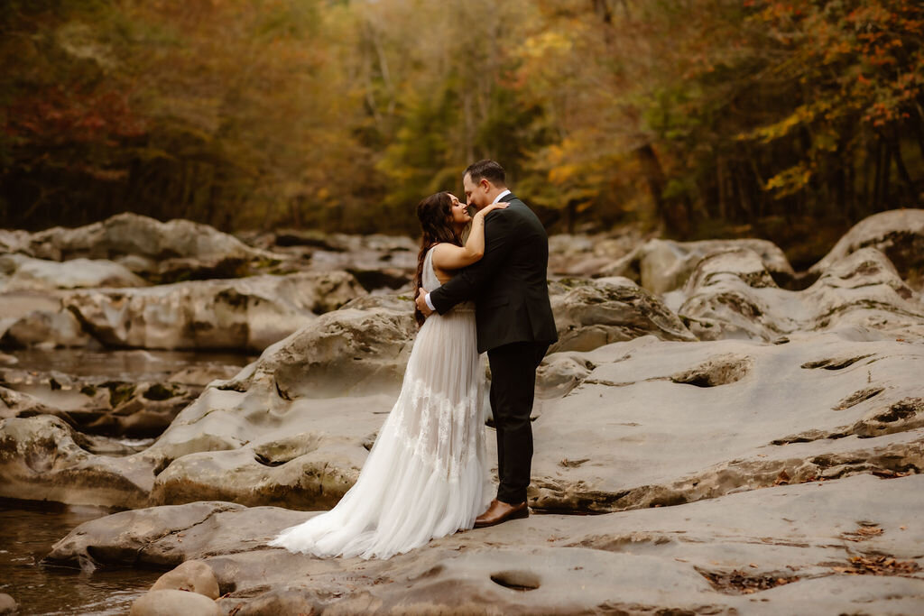 Bride and groom sharing a kiss beside the river at Greenbrier after their ceremony, surrounded by smooth rocks and fall foliage while eloping to Gatlinburg.
