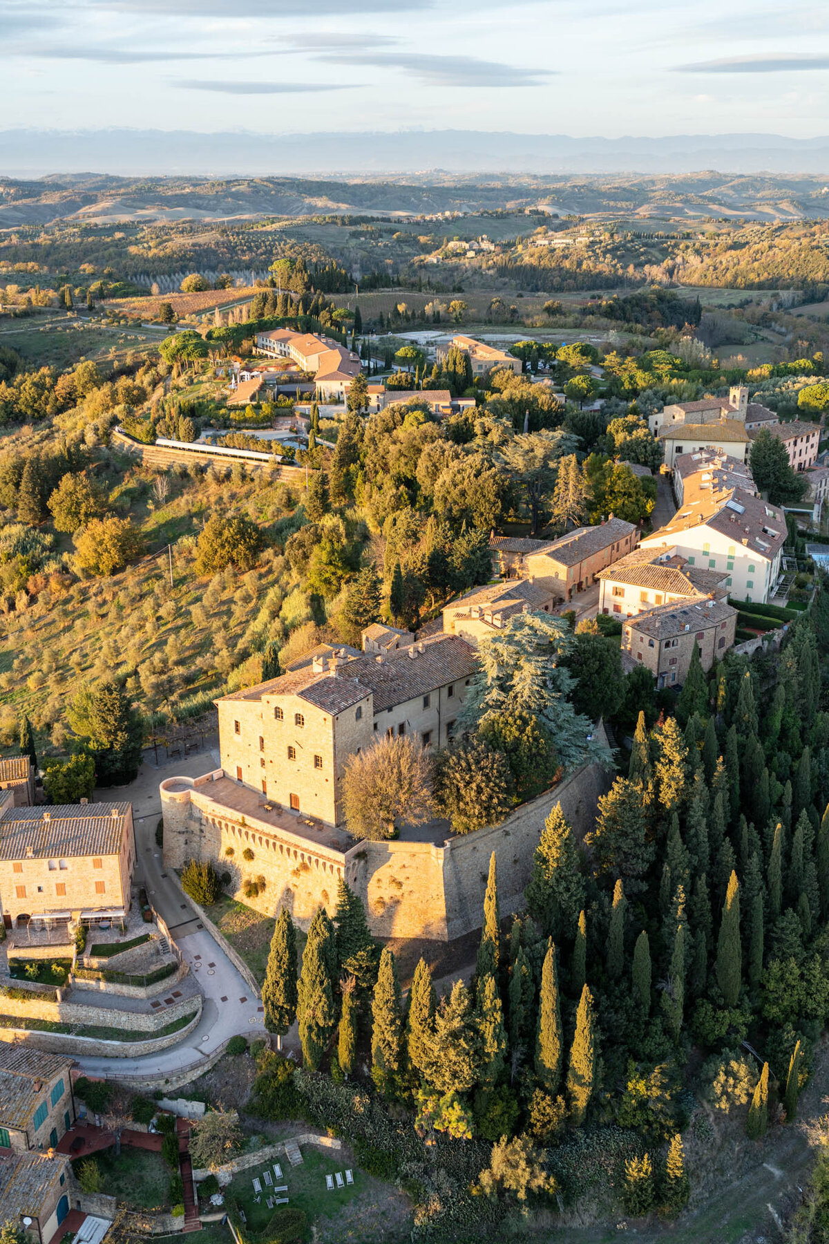 Aerial view of a Tuscan countryside with lush greenery, rolling hills, and a cluster of rustic buildings. The landscape features winding roads, tall cypress trees, and fields bathed in golden sunlight.