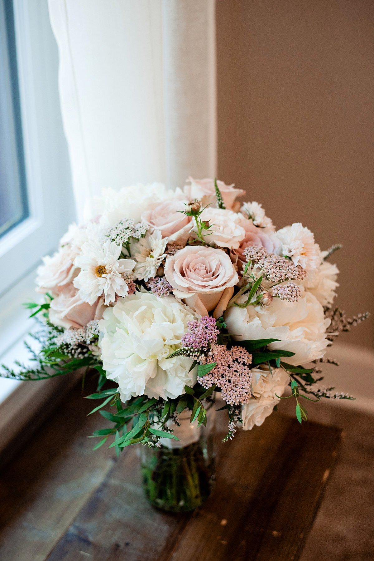 Bridal bouquet of white peonies, blush roses, white dahlias and pink rice flower accented with greenery sits in a glass vase on a window sill.