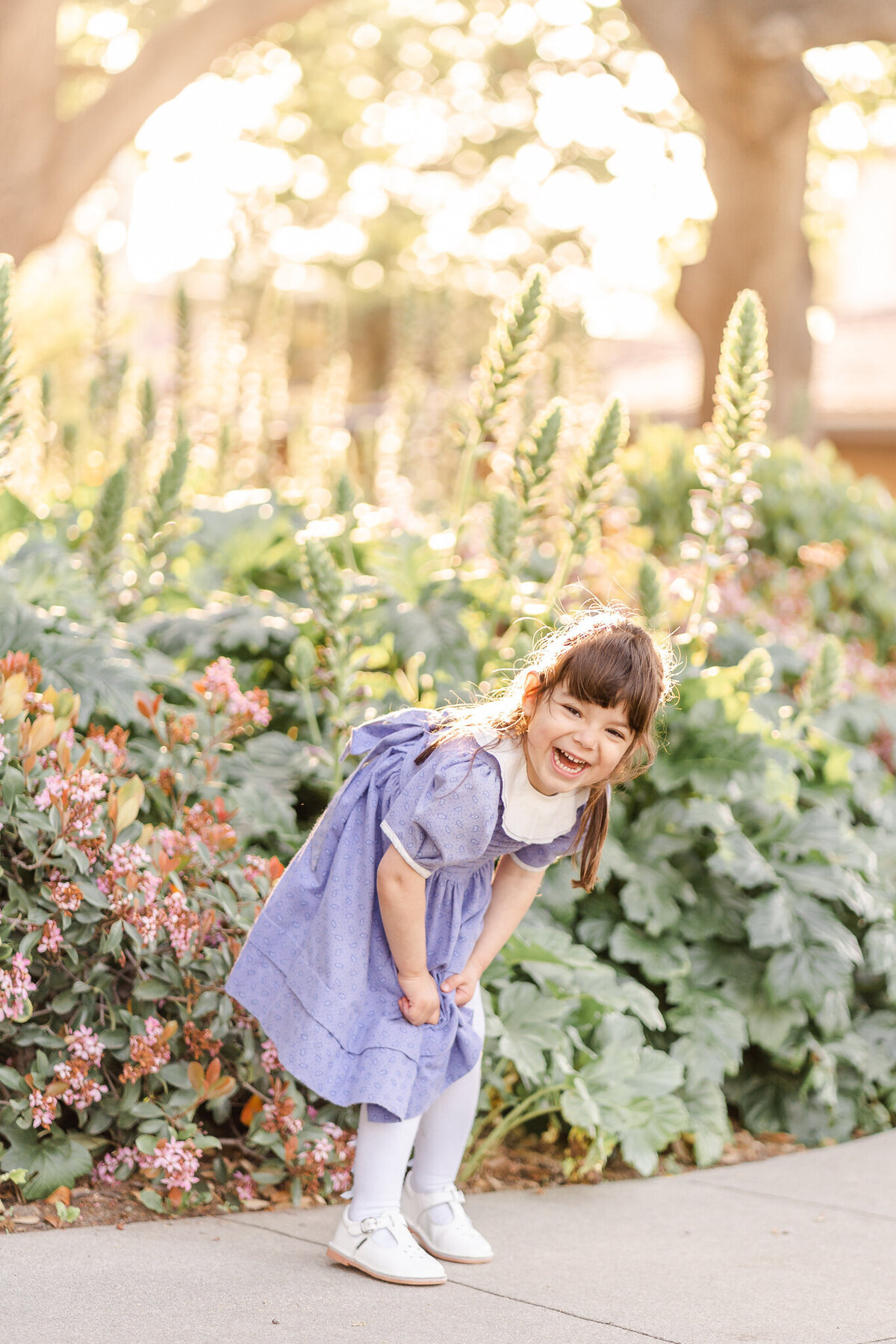 little girl in a lavender dress laughing big in front of a beautiful flower garden