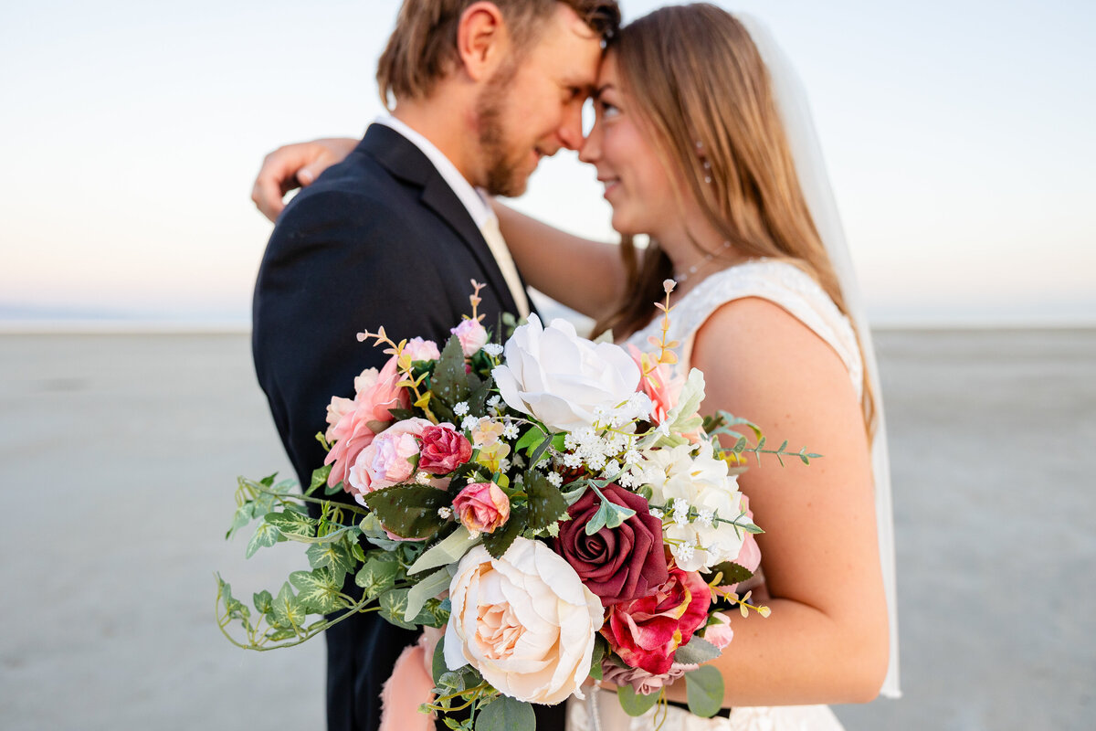 Bride and groom embrace closely, with a vibrant bouquet of roses and greenery in the foreground.