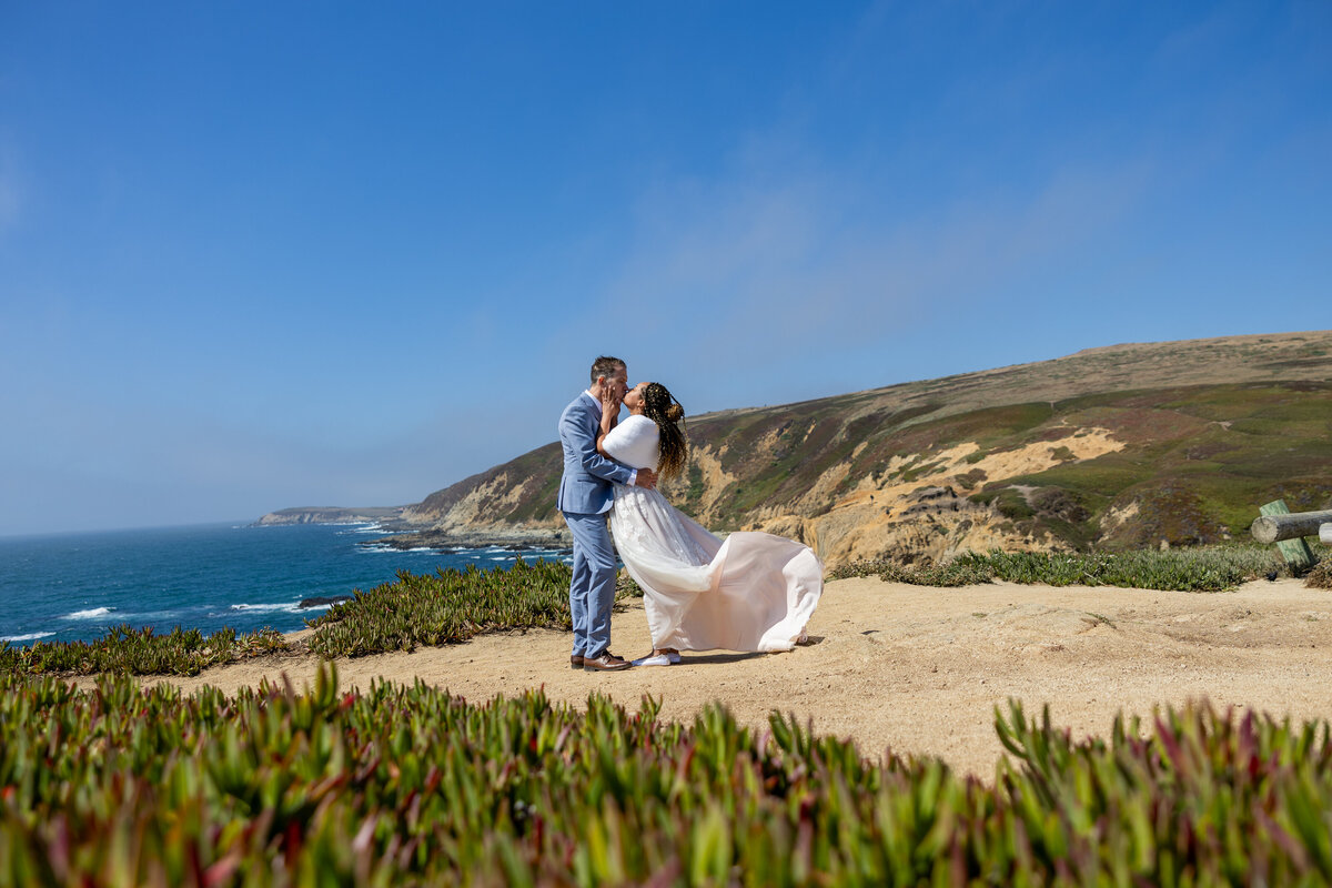 Bride and groom kiss in front of the ocean in Monterey.  Wind lifts the bottom of bride's dress up in a whimsical way. Photo by philippe studio pro, sacramento wedding photographer.