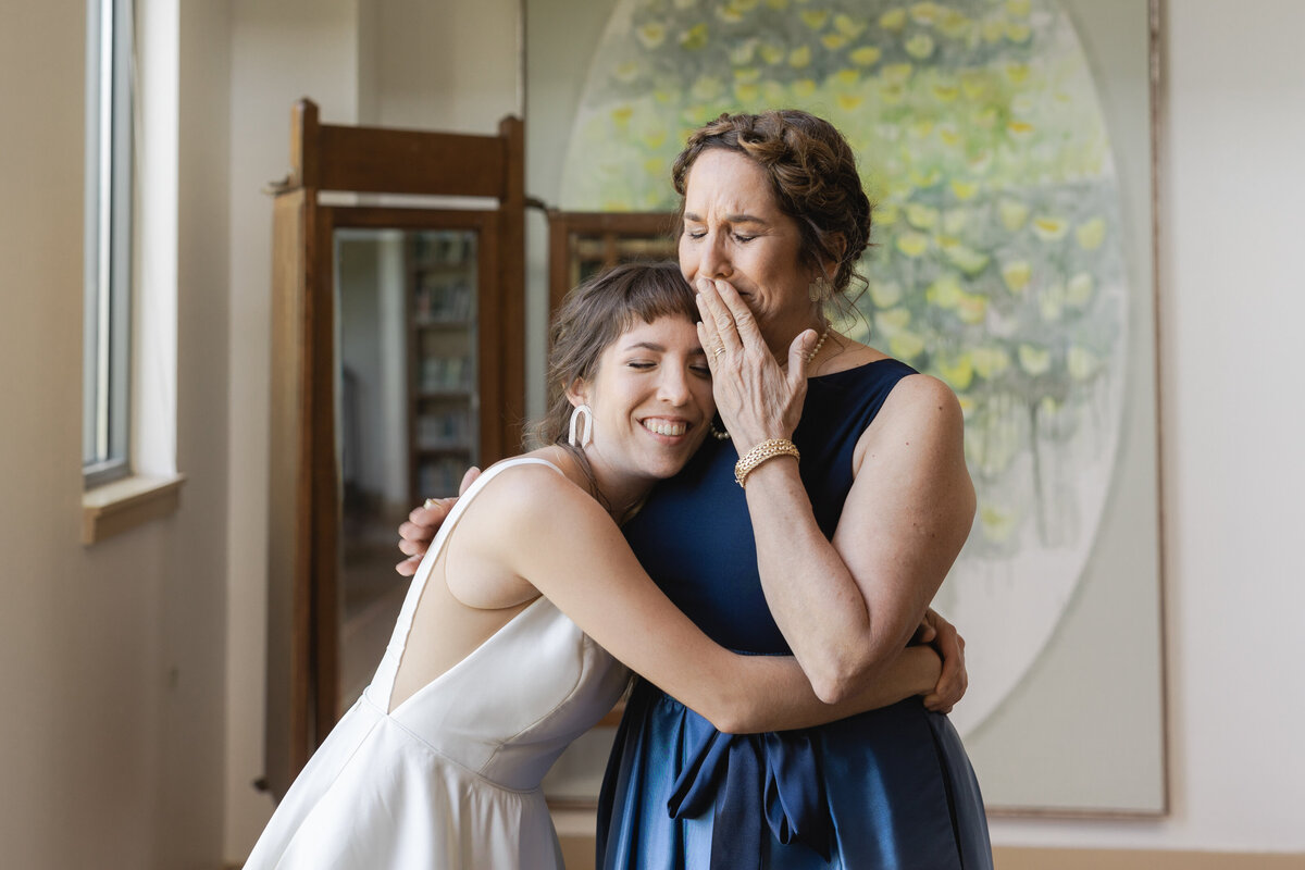 A candid shot of a bride hugging her mother after seeing her for the first time on her wedding day at the Lady Bird Johnson Wildflower Center in Austin, Texas. The bride is wearing a sleeveless, white dress and is hugging her mother closely. The mother is holding back tears and is wearing an elegant, blue dress. They both are in front of a large mirror and painting.