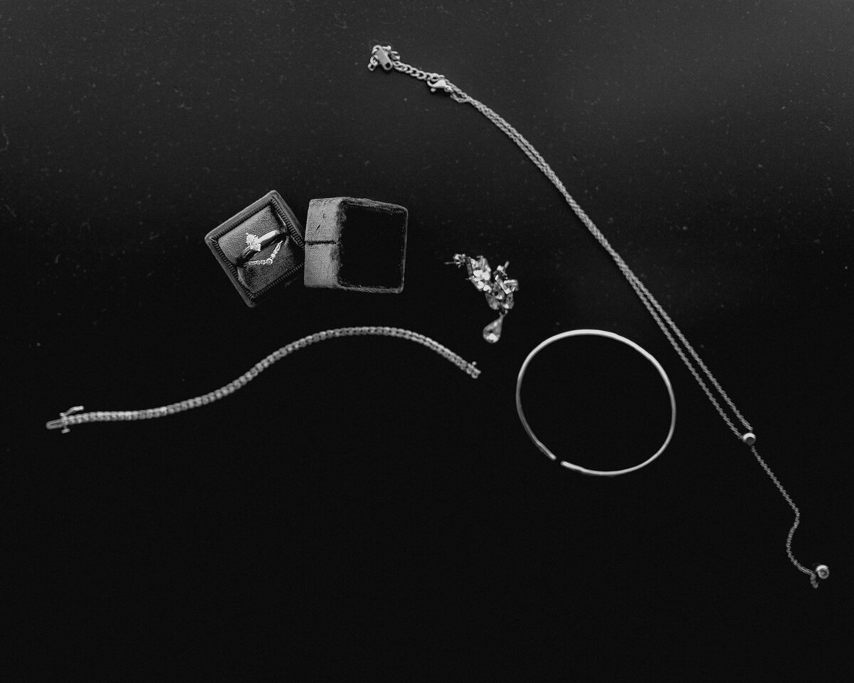 Black and white close-up of bridal jewelry, including a ring in a velvet box, bracelet, earrings, and a delicate necklace, placed on a dark background.