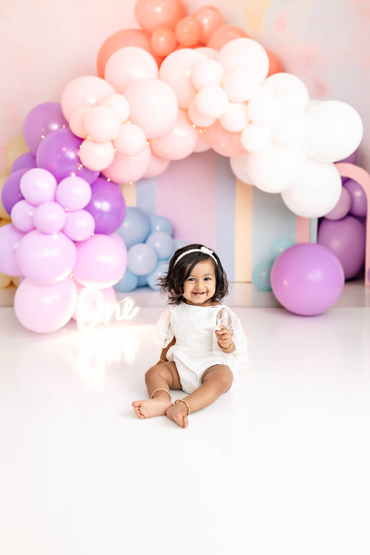 A smiling toddler in a white outfit sits on the floor in front of a pastel balloon arch with pink, purple, and white balloons. The background is bright, and the word "One" is visible in illuminated letters.