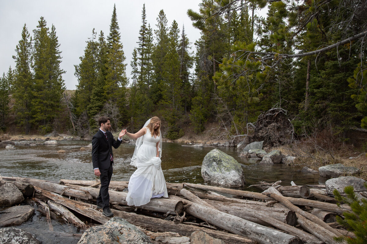 A groom helps his bride walk across logs in a river on their elopement day.