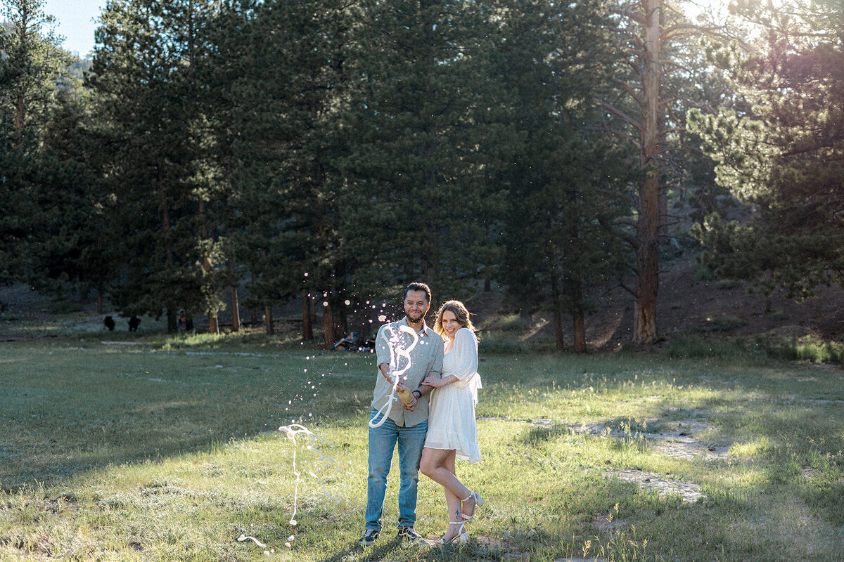 A joyful moment of the couple standing together in a grassy field as champagne bursts from a bottle, creating a celebratory spray.