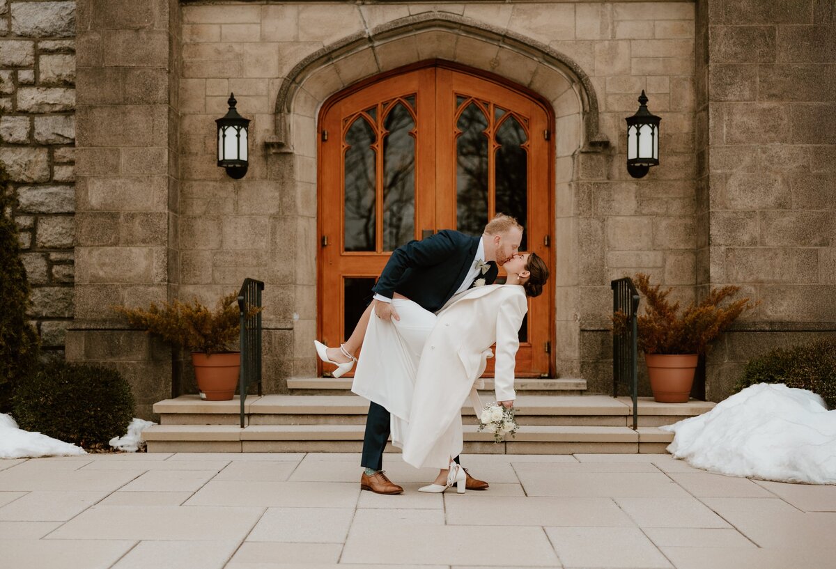 A newlywed couple shares a romantic dip and kiss in front of a stone church with wooden double doors in the Hudson Valley. The groom wears a navy suit, and the bride wears a white coat and heels, holding a bouquet. Patches of snow surround the steps, adding to the winter wedding charm.