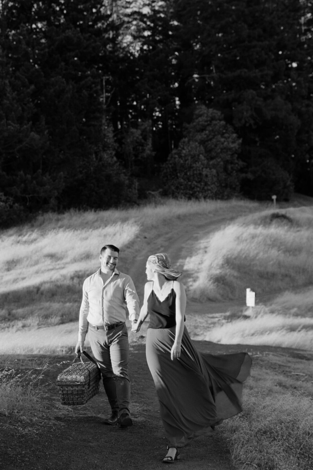 Man carries a basket and follows his jubilant romantic partner through the meadows.