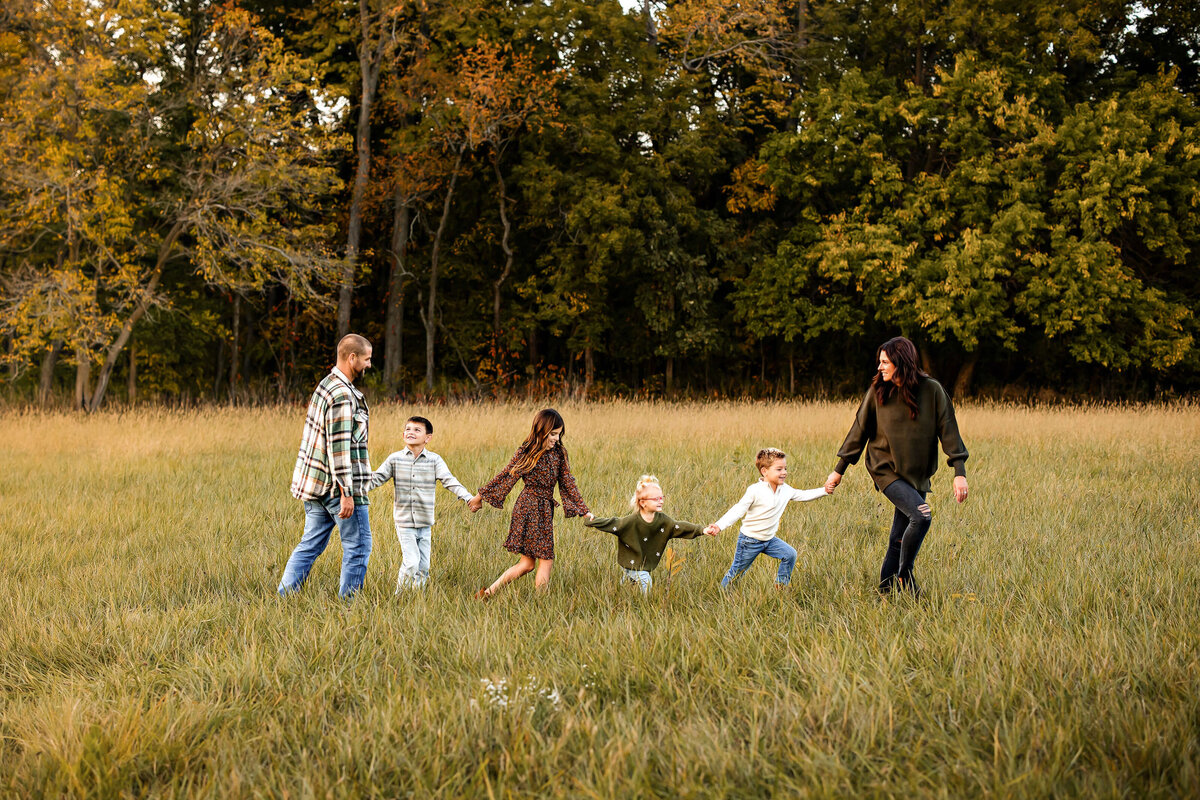A family of six holding hands and walking in a grassy field under tall trees with autumn leaves. The parents are on either end, and the four children are in between, smiling and enjoying the outdoors.