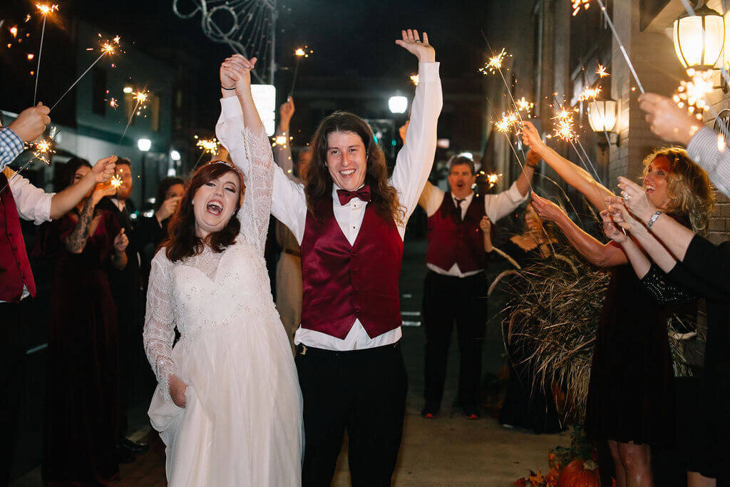 A bride and groom celebrate as they walk hand-in-hand through a cheering crowd holding sparklers outside at night. The couple smiles joyfully, surrounded by friends and family lined along the sidewalk.